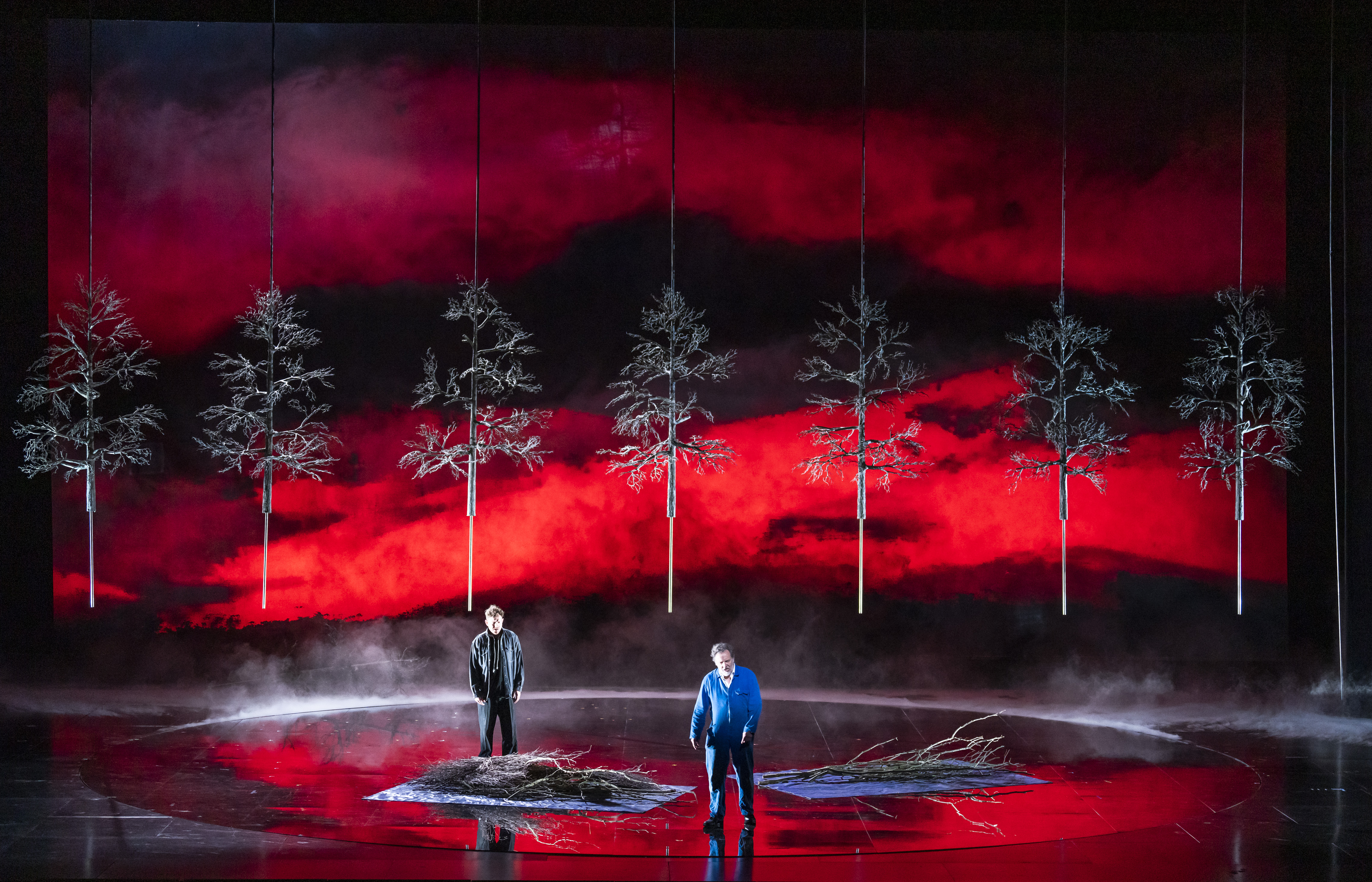 Two performers stand on a darkened stage with a deep red background with bare trees.