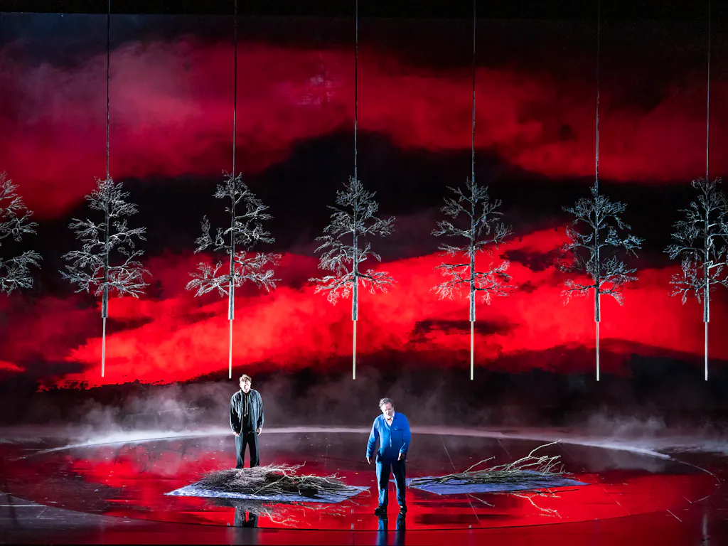 Two performers stand on a darkened stage with a deep red background with bare trees.