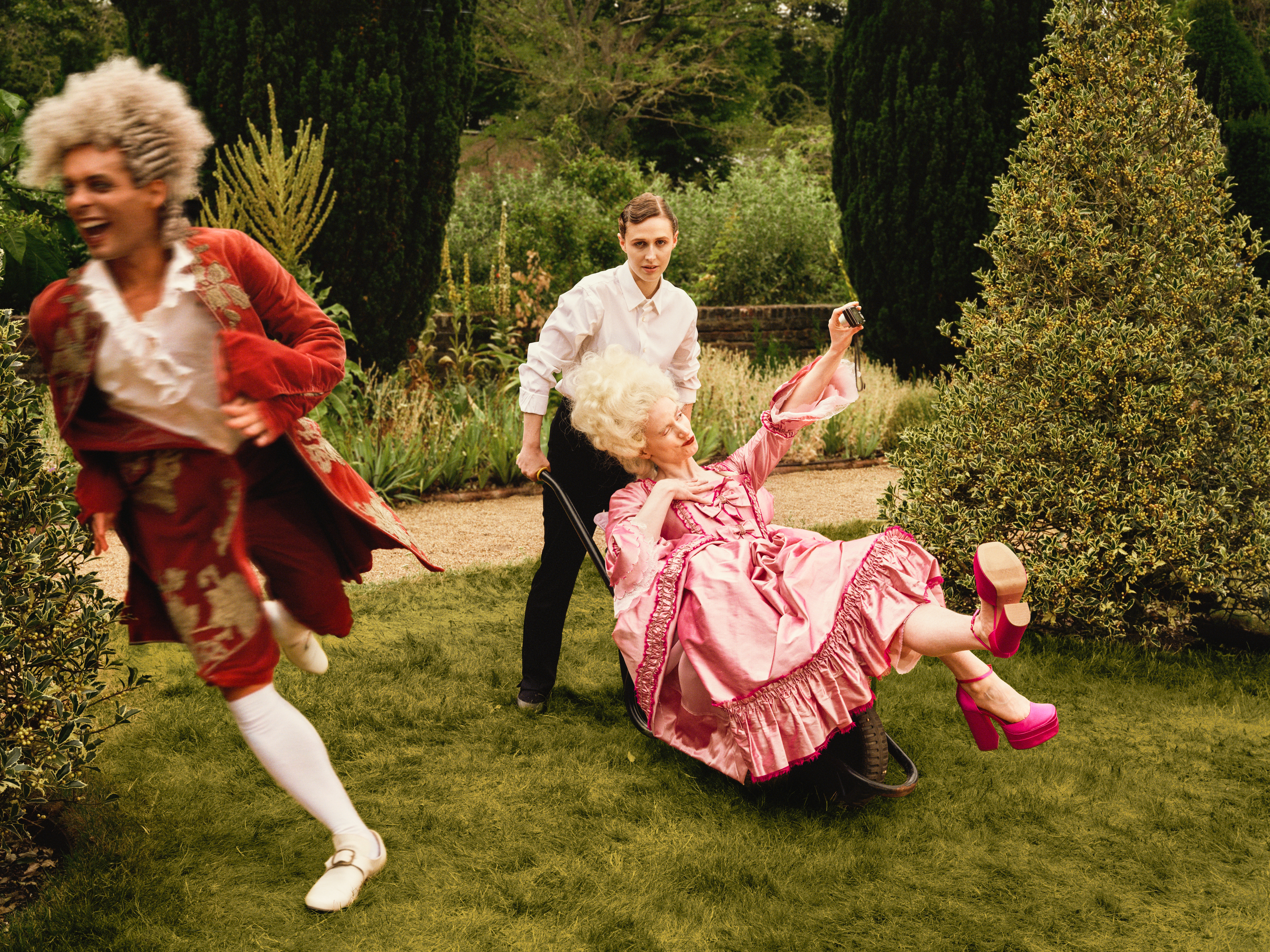 Three performers in a formal garden: a woman in a black and white maiden costume pushes a performer in an elaborate pink period costume seated on a garden wheelbarrow, while another performer in a red period costume runs past in the foreground. (4 of 4)