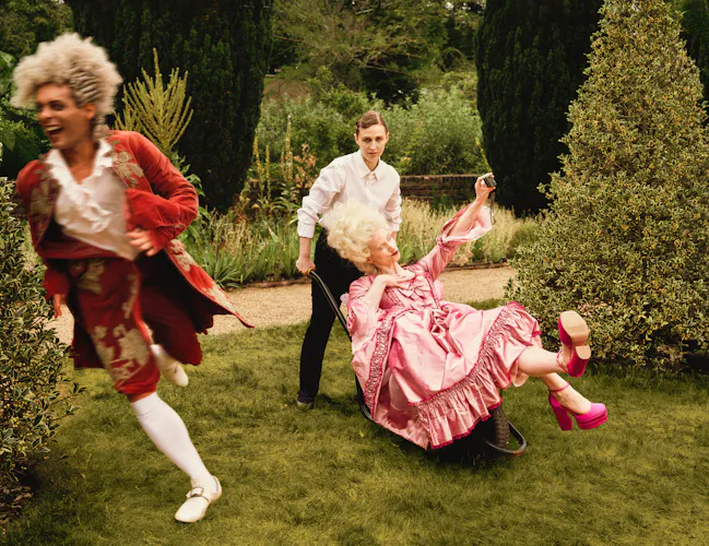 Three performers in a formal garden: a woman in a black and white maiden costume pushes a performer in an elaborate pink period costume seated on a garden wheelbarrow, while another performer in a red period costume runs past in the foreground. (4 of 4)