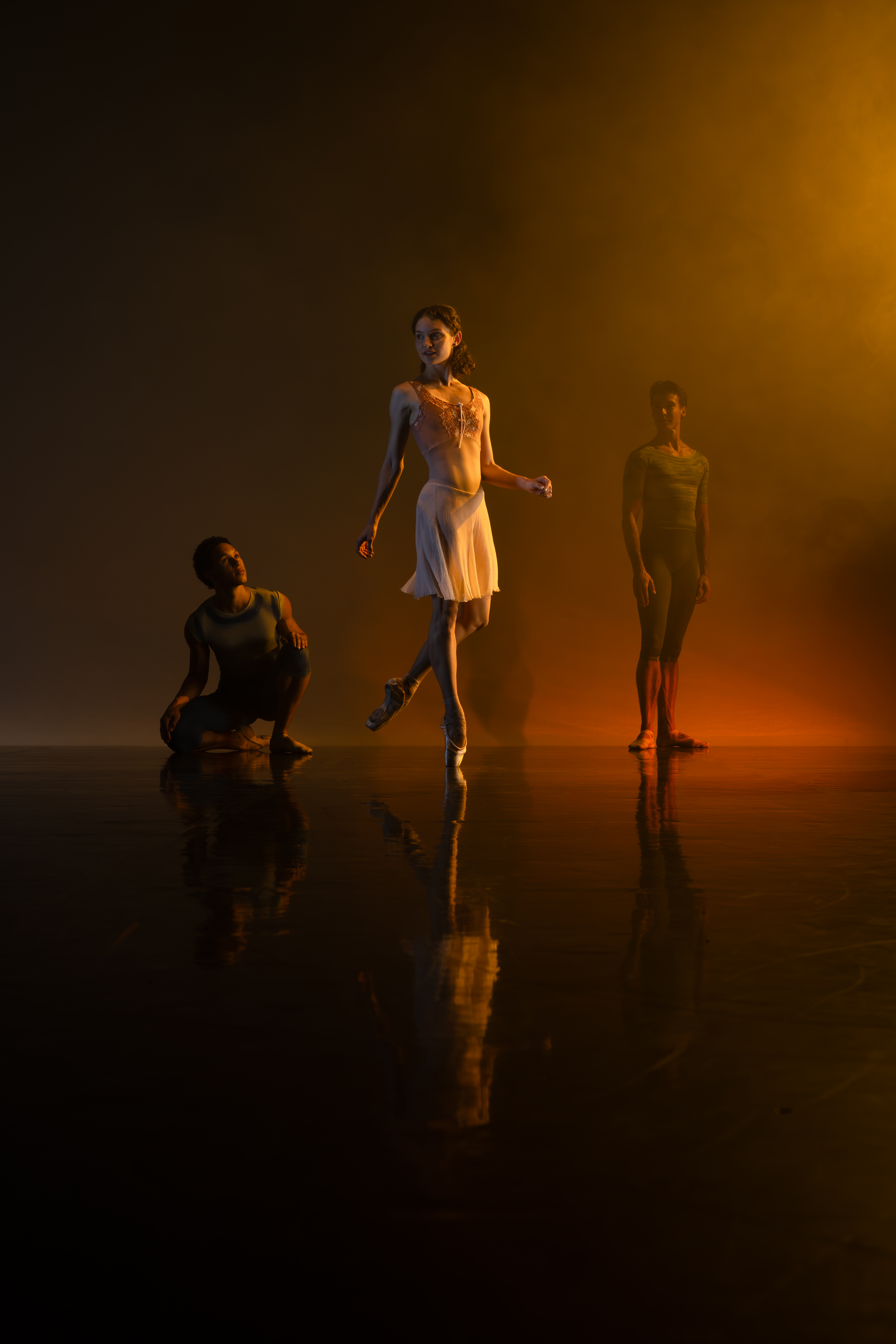 Three dancers stand on a darkened stage. An orange light shines on them from above. One dancer in a white dress stands in the middle and slightly in front. The other two dancers wear dark outfits.