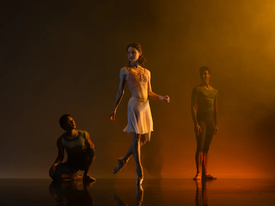 Three dancers stand on a darkened stage. An orange light shines on them from above. One dancer in a white dress stands in the middle and slightly in front. The other two dancers wear dark outfits.