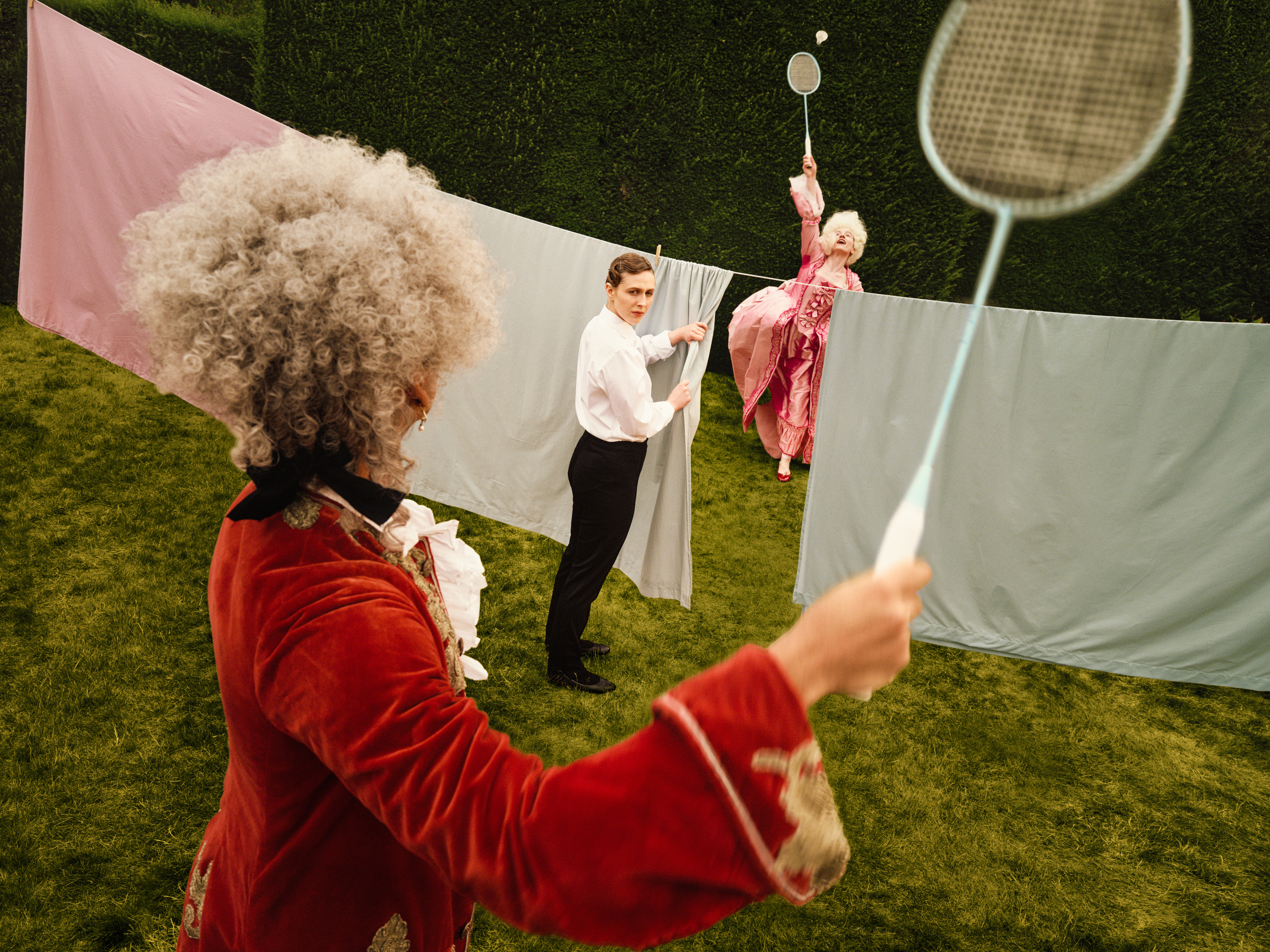 Two performers in elaborate period costumes playfully move through a hedge garden with sheets of fabric hung between them. In the foreground, a performer in a red velvet costume holds a badminton racket and is playing with a woman in a pink costume on the other side of the washing line. In the middle is a third person, representing Despina in Netia Jones’s Così fan tutte, dressed as a maid and hanging the laundry in the middle of the scene. (1 of 4)