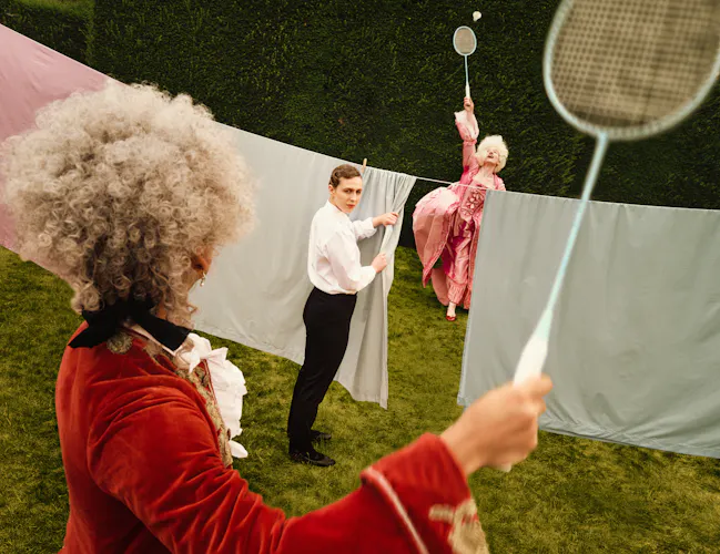 Two performers in elaborate period costumes playfully move through a hedge garden with sheets of fabric hung between them. In the foreground, a performer in a red velvet costume holds a badminton racket and is playing with a woman in a pink costume on the other side of the washing line. In the middle is a third person, representing Despina in Netia Jones’s Così fan tutte, dressed as a maid and hanging the laundry in the middle of the scene. (1 of 4)