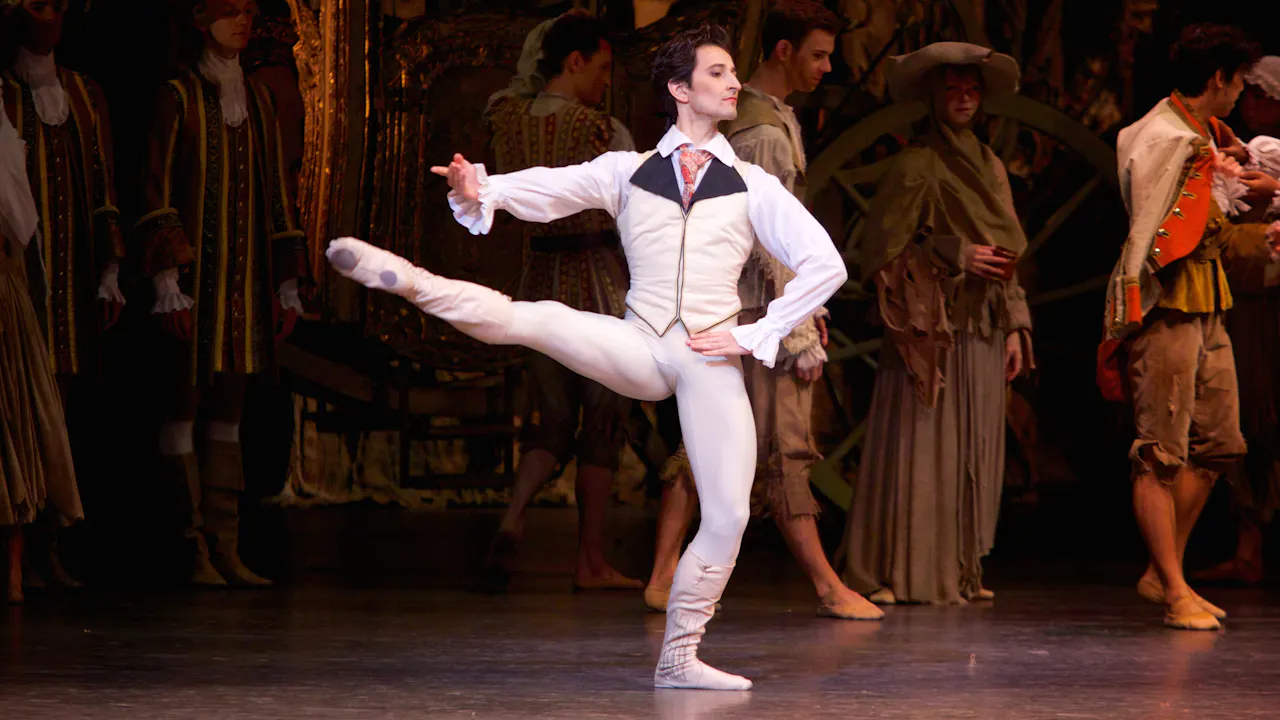 A dancer dressed as a Lord in a shirt and waistcoat balances on one leg in the centre of the stage