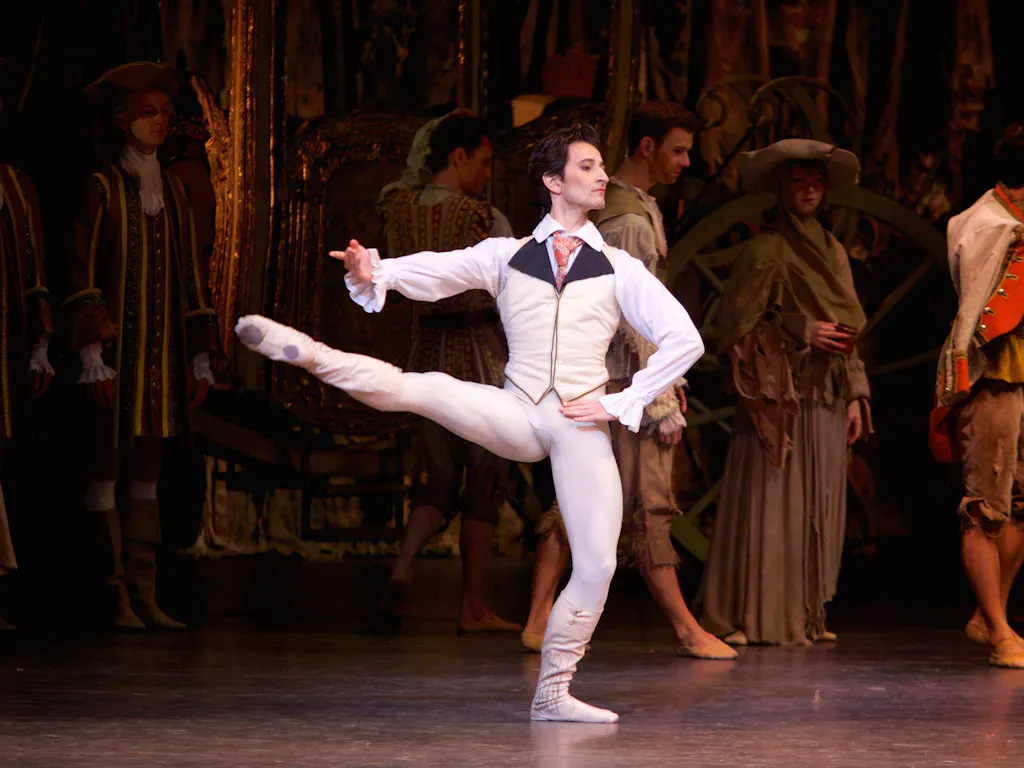 A dancer dressed as a Lord in a shirt and waistcoat balances on one leg in the centre of the stage