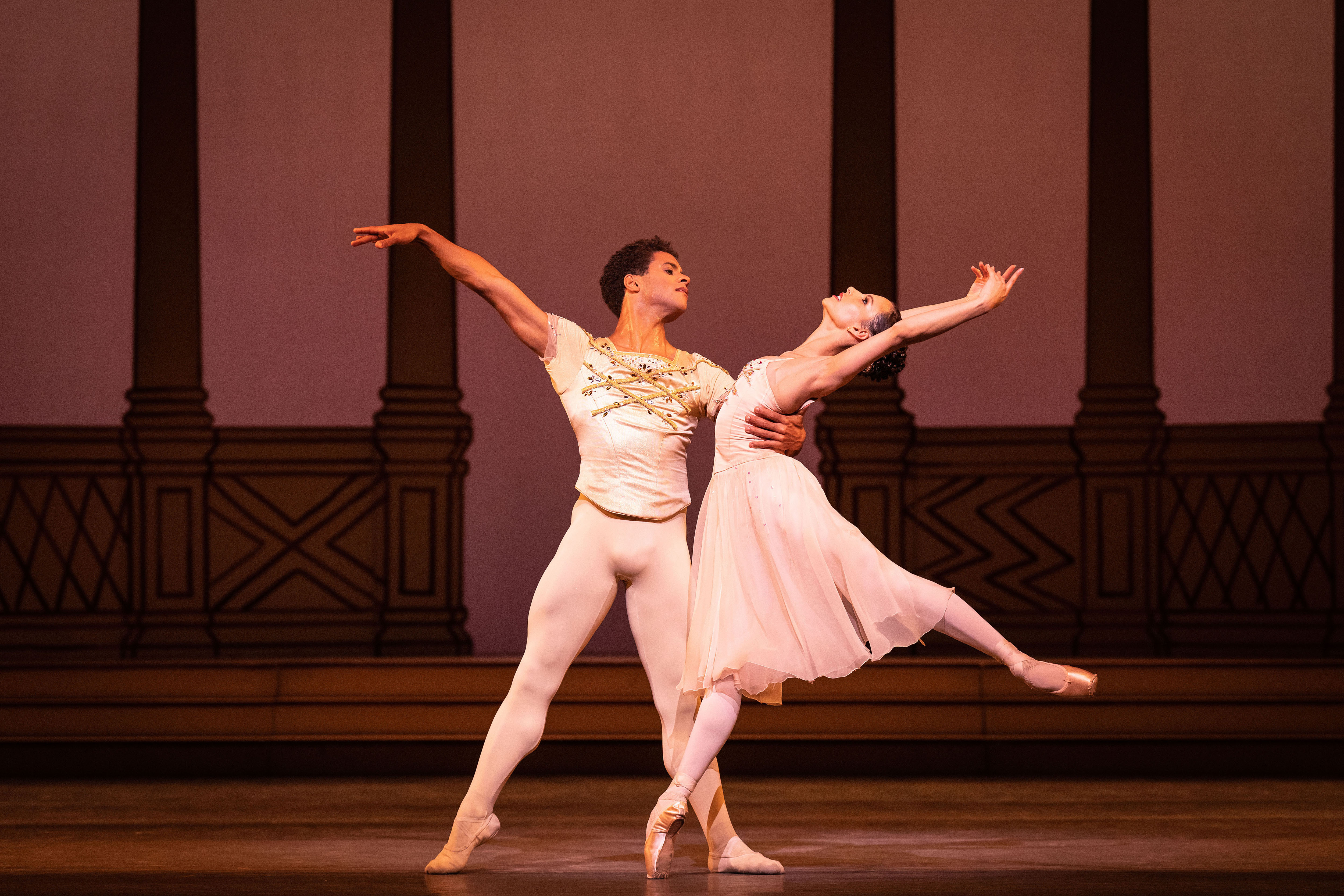 Two dancers perform on stage. They man wears white tights and a white tight, top. The woman wears a pink dress. Behind them columns can be seen. (1 of 8)