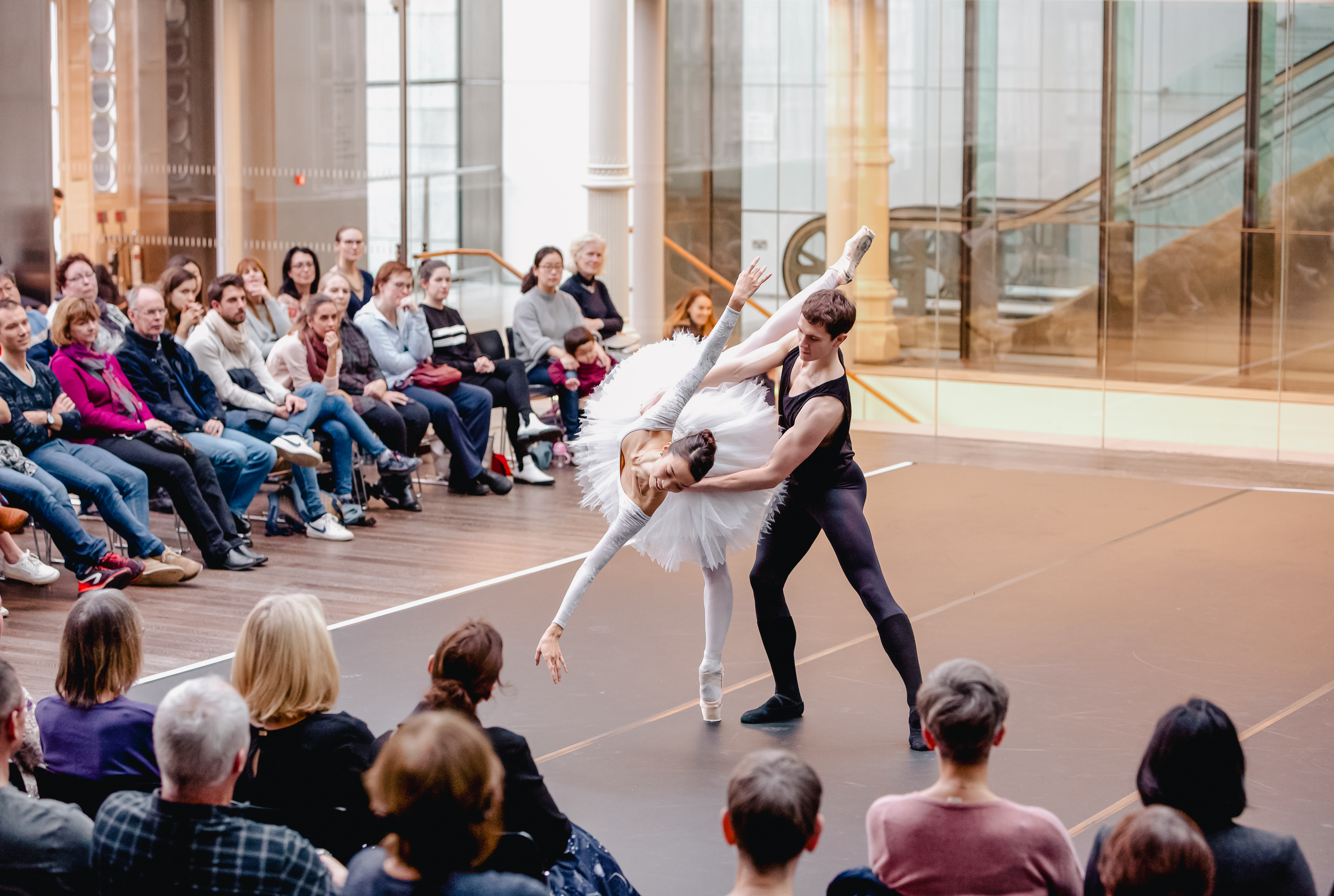 Two dancers appear in the Paul Hamlyn Hall. The man wears a black leggings and a black vest. The woman wears a white leotard and tutu. A crowd sit around them.