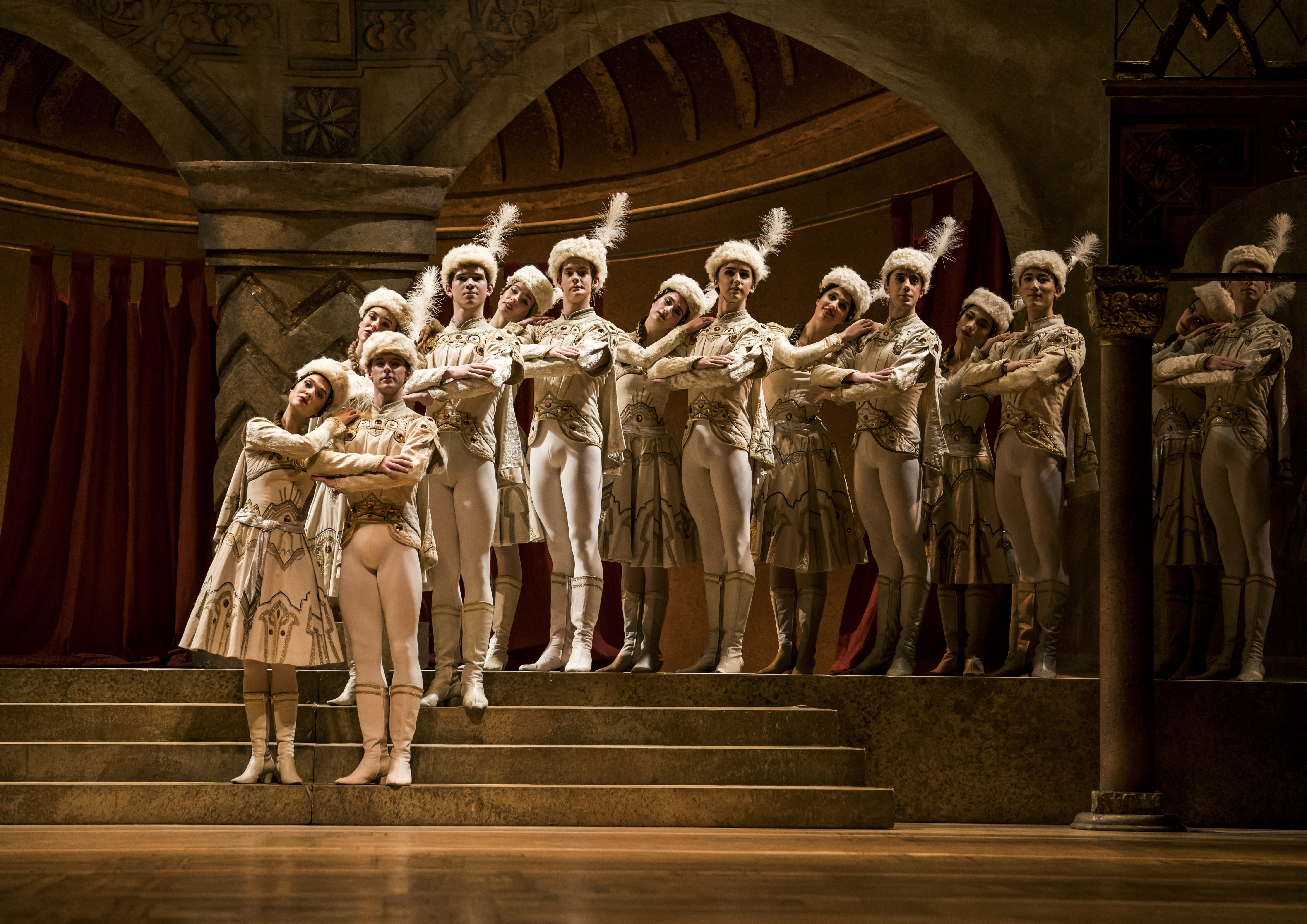 A group of dancers stand on a set of steps on stage, behind them ornate are ornate archways. They wear decorative formal white outfits and have fur hats with white feathers. 