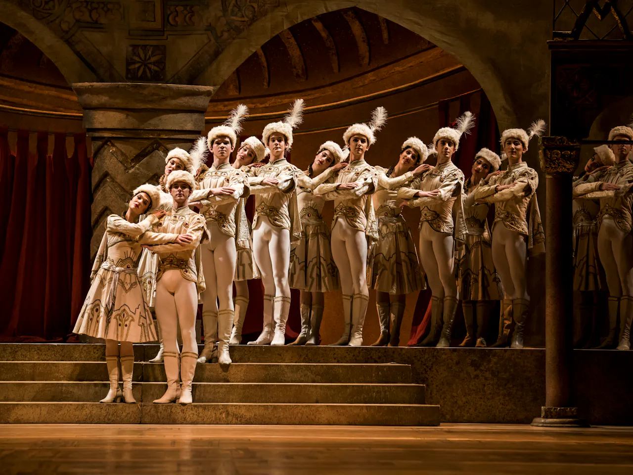 A group of dancers stand on a set of steps on stage, behind them ornate are ornate archways. They wear decorative formal white outfits and have fur hats with white feathers.