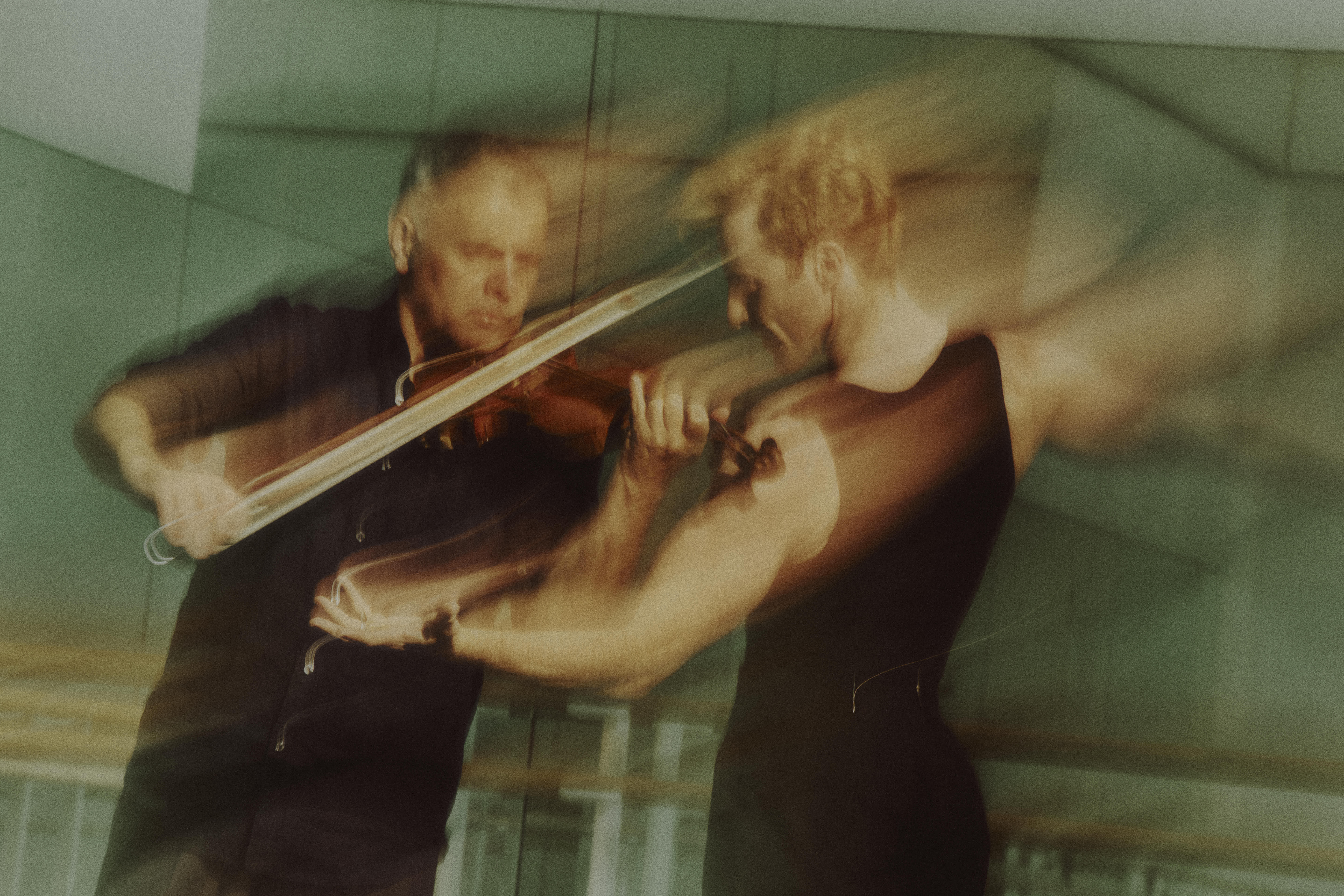 A blurred image of Steven McRae and Vasko Vassilev in rehearsal. Both wear black, Vassilev plays his violin while McRae moves in front with arms partially outstretched