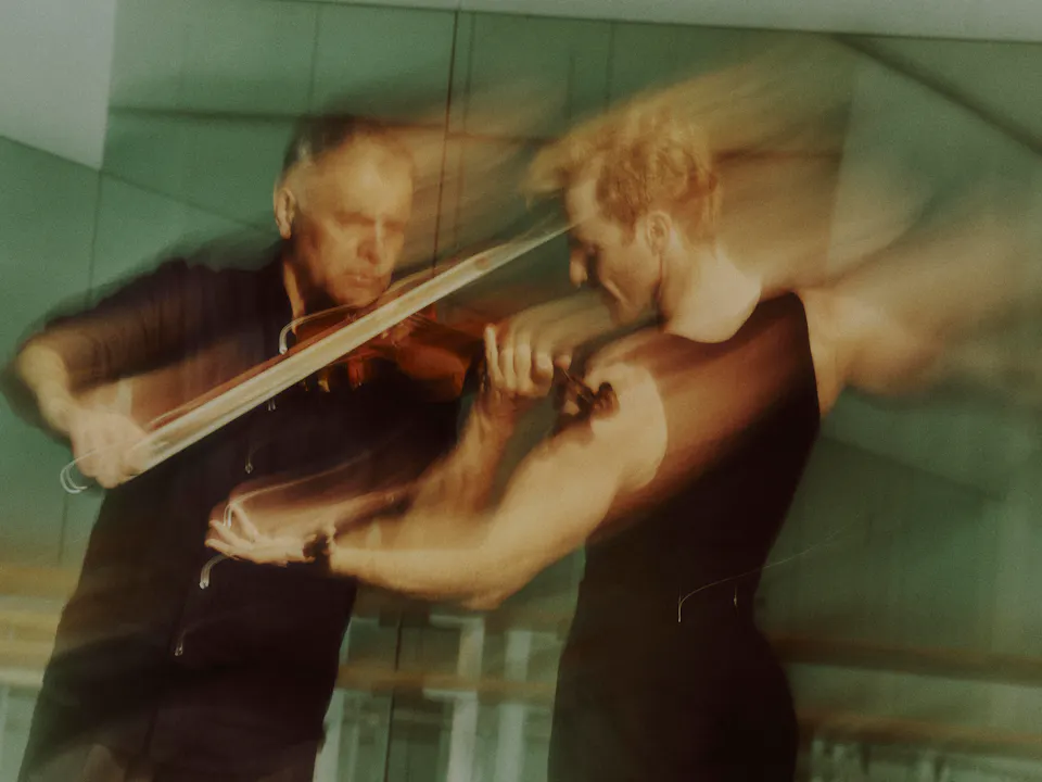 A blurred image of Steven McRae and Vasko Vassilev in rehearsal. Both wear black, Vassilev plays his violin while McRae moves in front with arms partially outstretched