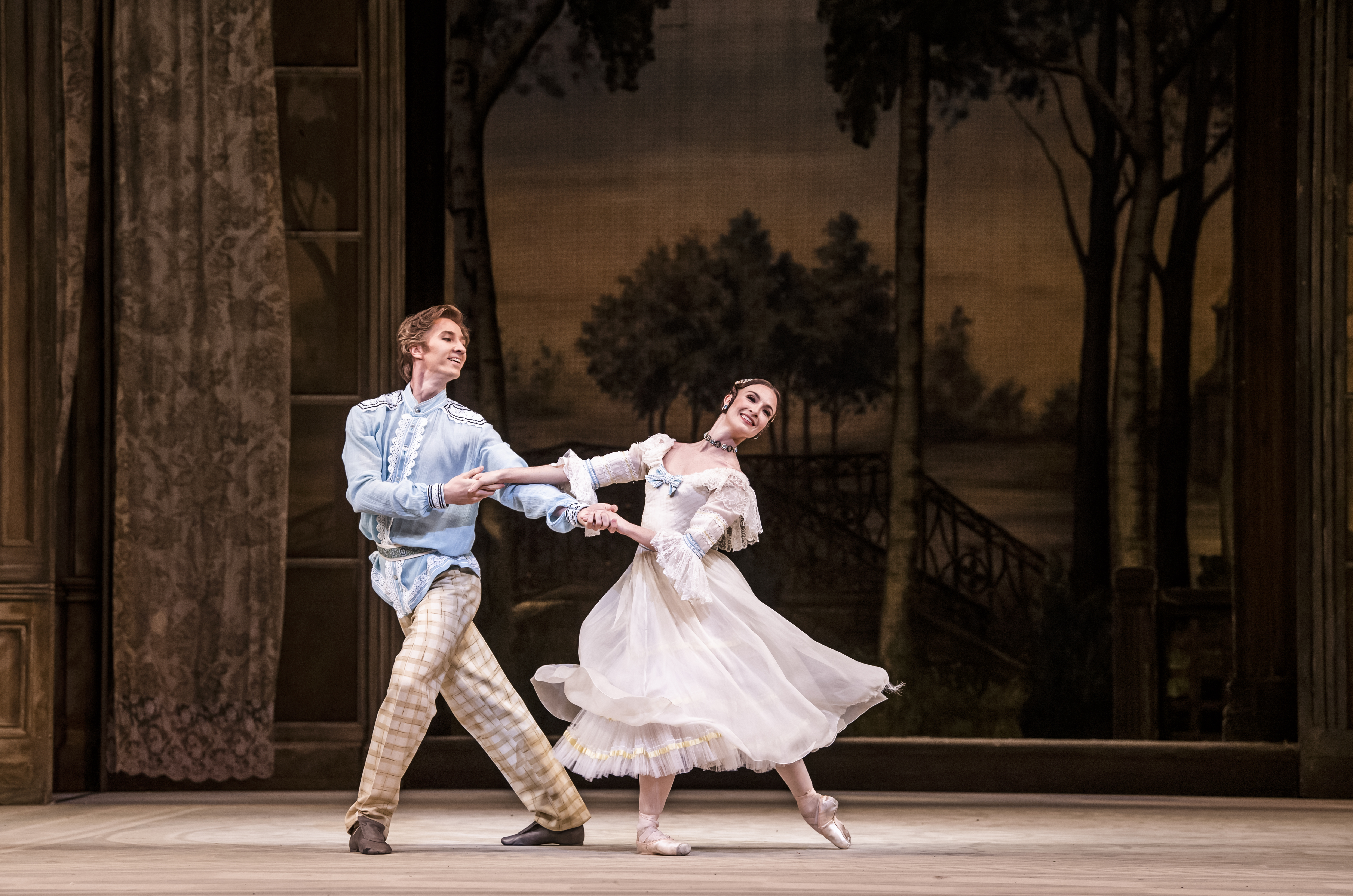 Two dancers perform on stage, behind them is a backdrop with a woodlands and fields. The man wears a blue tunic with pattered trousers, the woman a flowing white dress. (3 of 3)