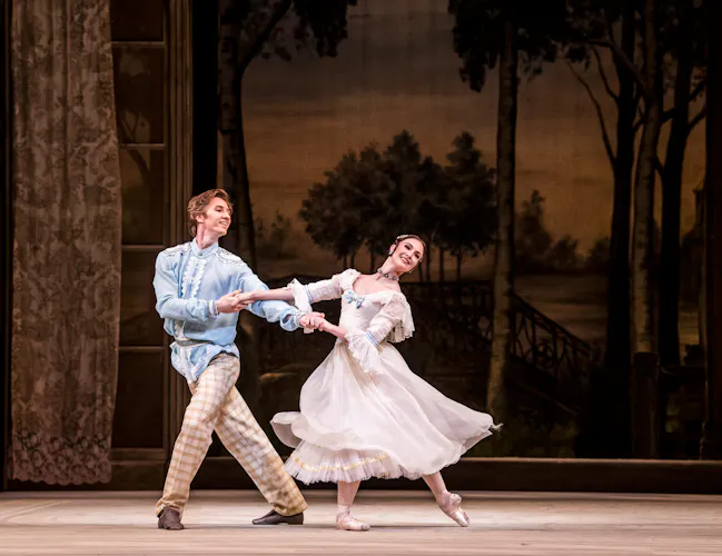 Two dancers perform on stage, behind them is a backdrop with a woodlands and fields. The man wears a blue tunic with pattered trousers, the woman a flowing white dress. (3 of 3)