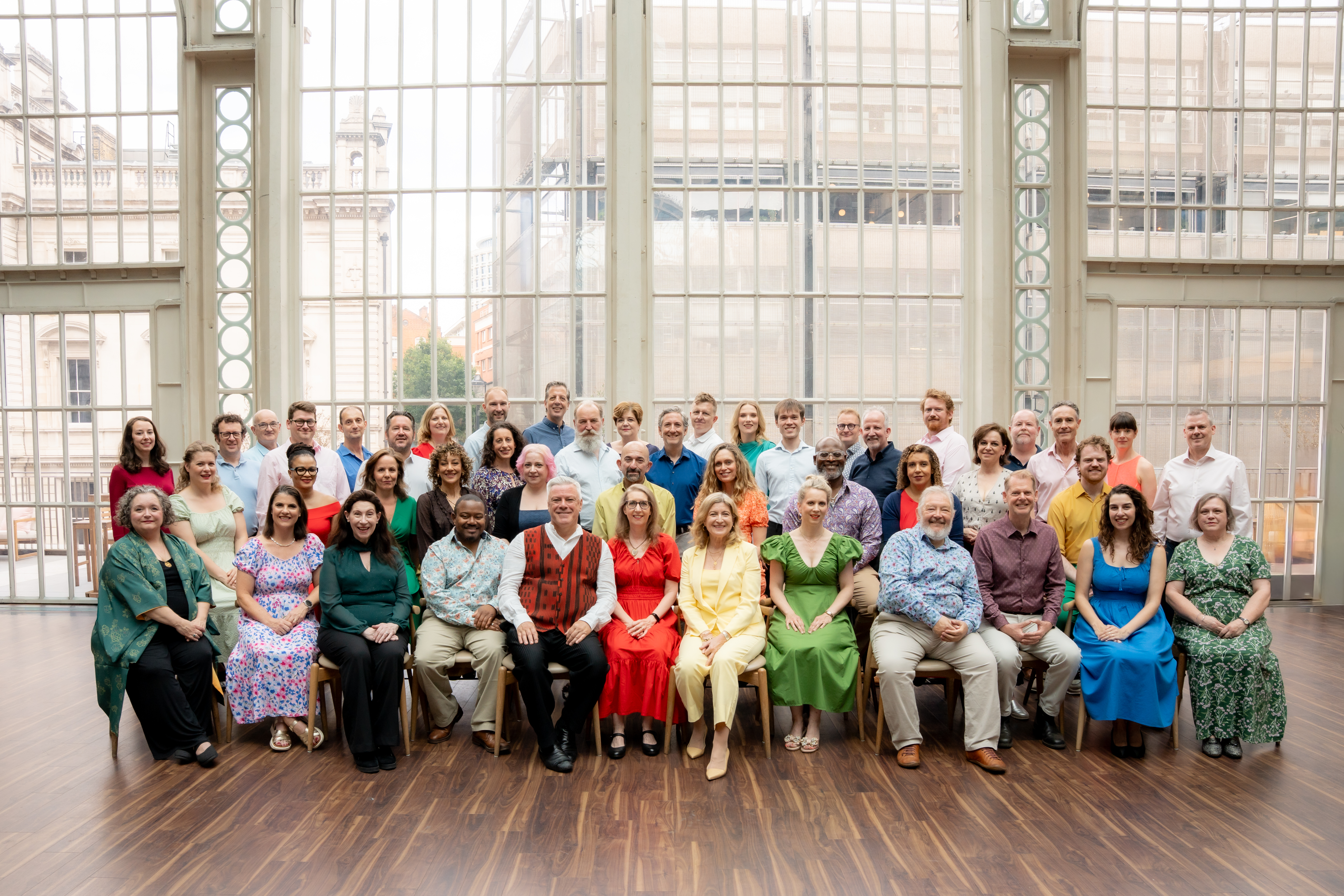 The Royal Opera chorus sit in the Paul Hamlyn Hall. They wear colourful outfits and sit in front of the glass facade.