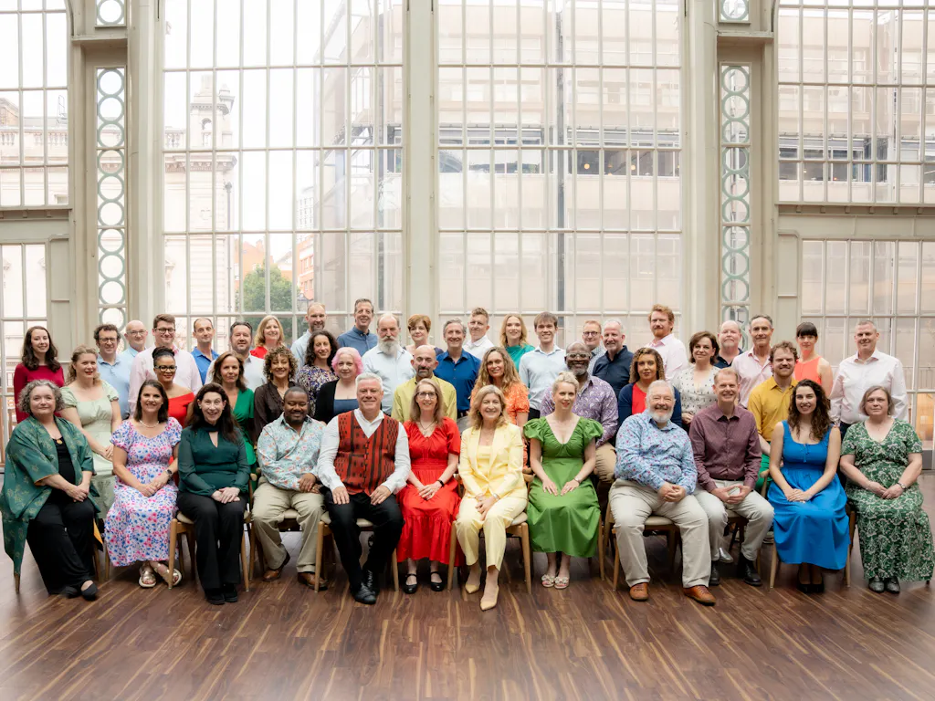 The Royal Opera chorus sit in the Paul Hamlyn Hall. They wear colourful outfits and sit in front of the glass facade.