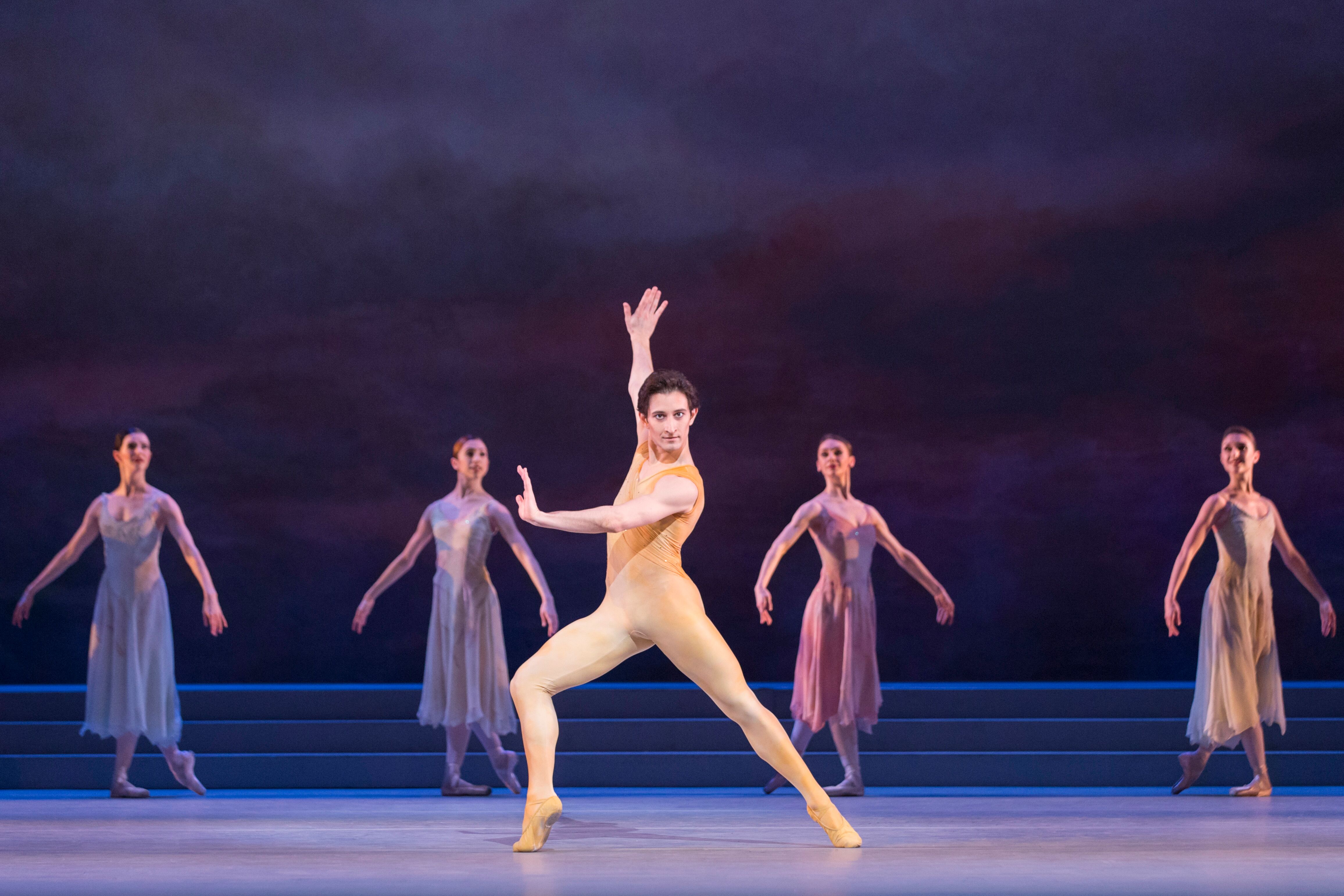 A male dancer in a nude leotard stands with arms and legs outstretched in the centre of the stage. Behind him are four female dancers in long purple dresses, standing in classical pose. (3 of 4)
