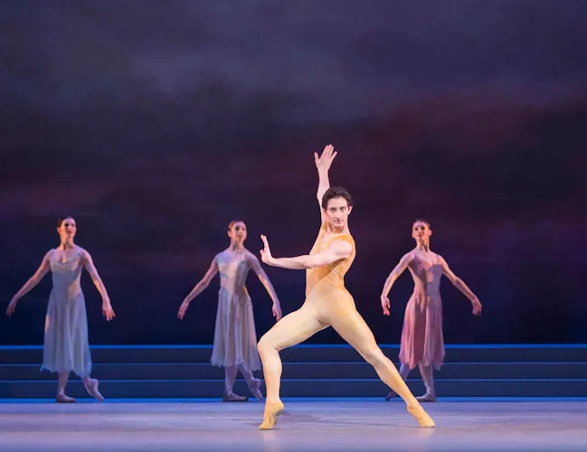 A male dancer in a nude leotard stands with arms and legs outstretched in the centre of the stage. Behind him are four female dancers in long purple dresses, standing in classical pose. (3 of 4)