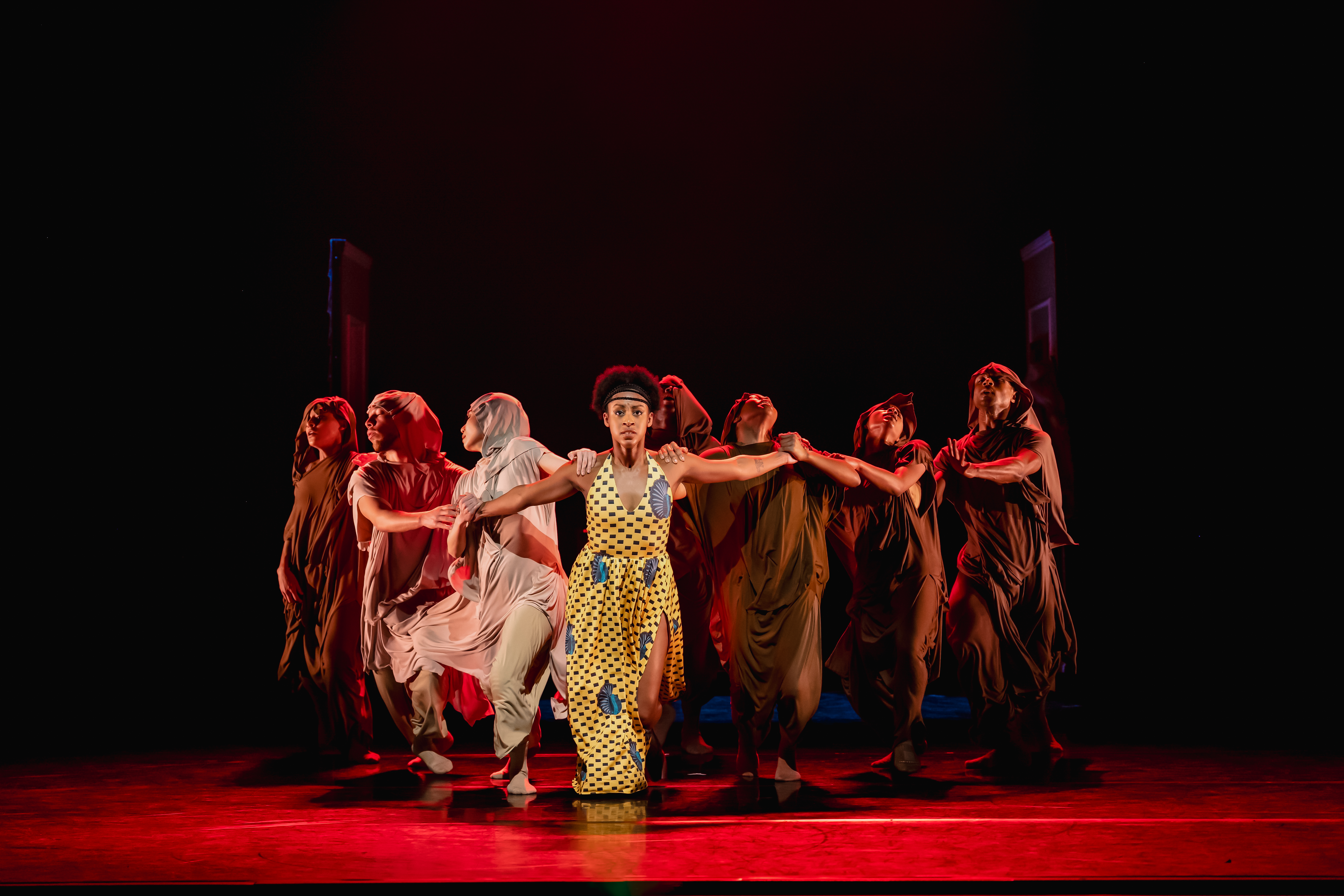 A woman in a yellow dress stands centre stage. An anonymous group of dancers hold her arms back, as if in a chain. 