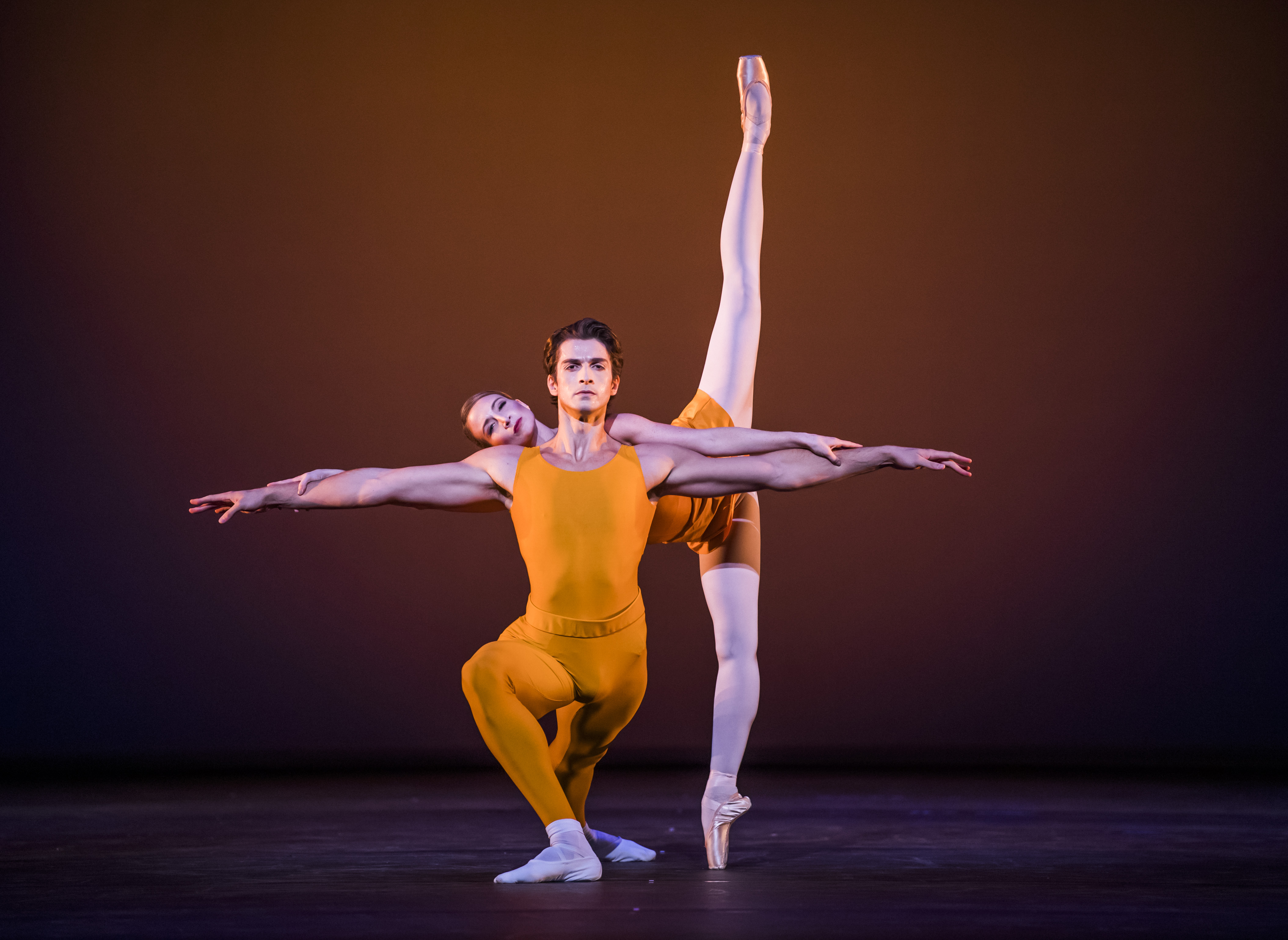 Two dancers wearing yellow leotards appear on stage. The dancer in front crouches with one knee bent in front of him and arms outstretched to the side. Behind him the other dancer has her legs in the splits in a vertical line from the floor, she balances her body on the man's back.