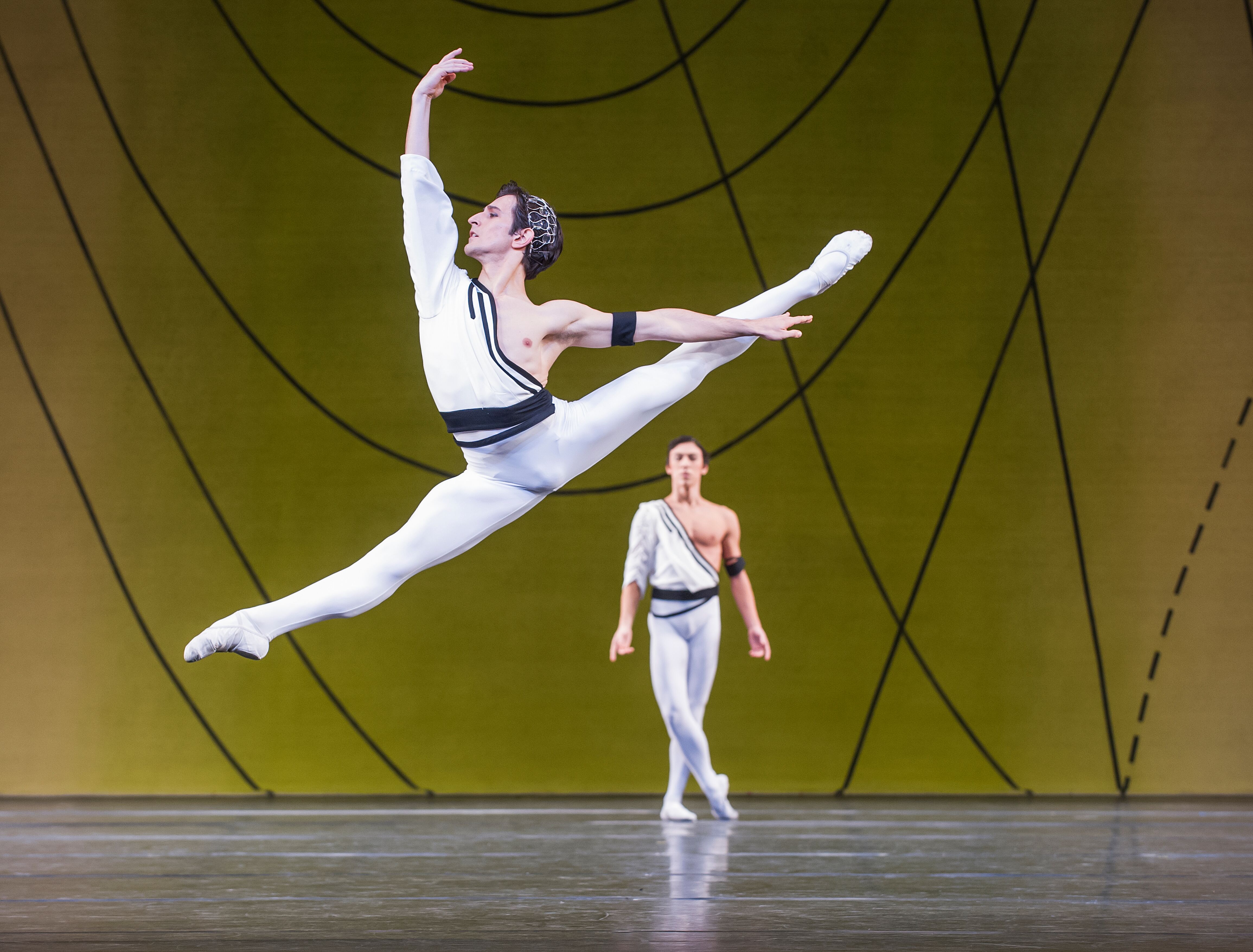 A male dancer wearing white tights and a black and white shirt leaps across the stage. Behind him is another male dancer in the same outfit, standing with his legs crossed. (2 of 4)