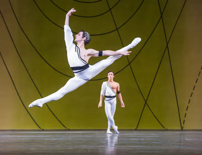 A male dancer wearing white tights and a black and white shirt leaps across the stage. Behind him is another male dancer in the same outfit, standing with his legs crossed. (2 of 4)