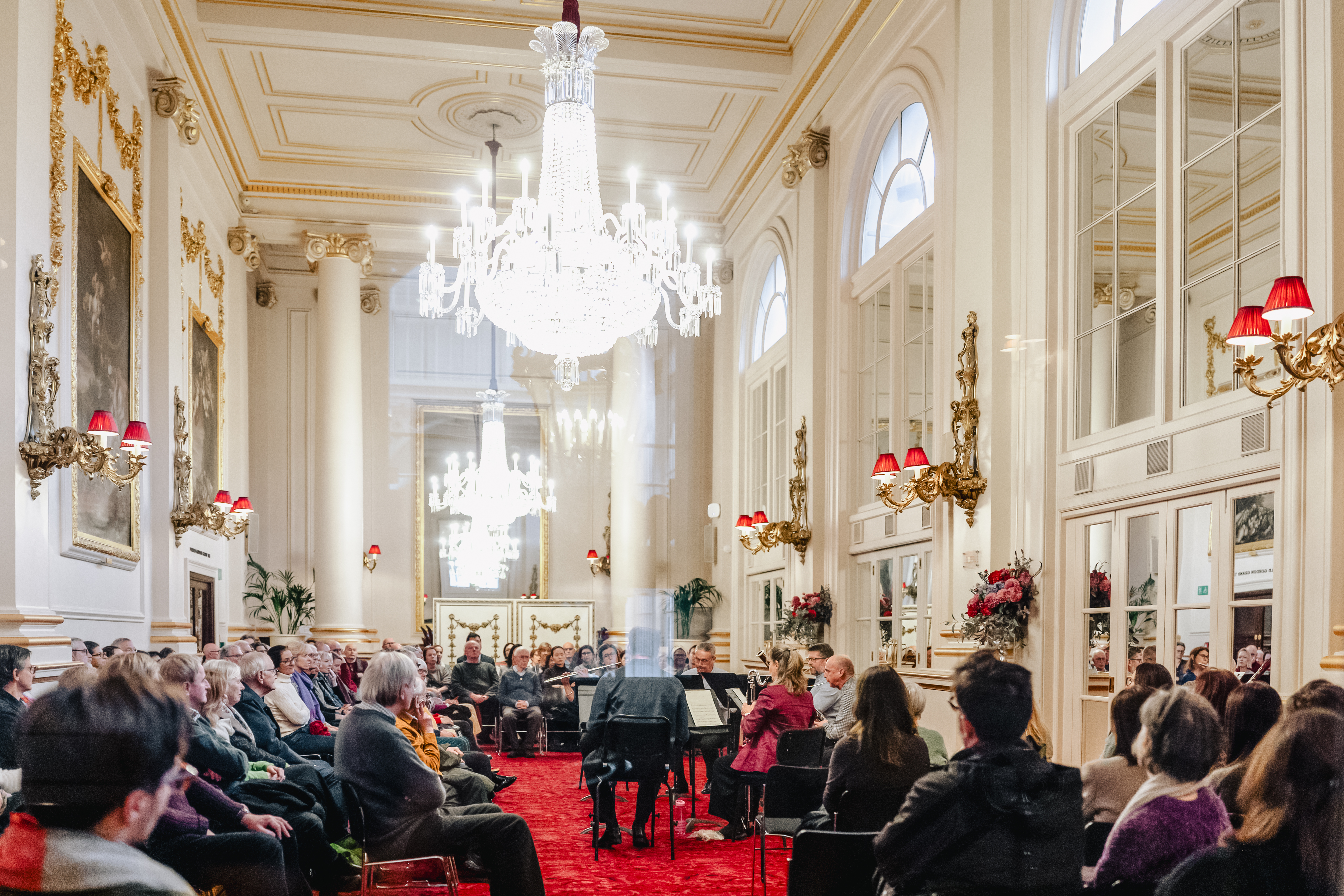 Many people sit around a small group of musicians in a brightly lit ornate room with chandeliers, oil paintings and mirrors. 