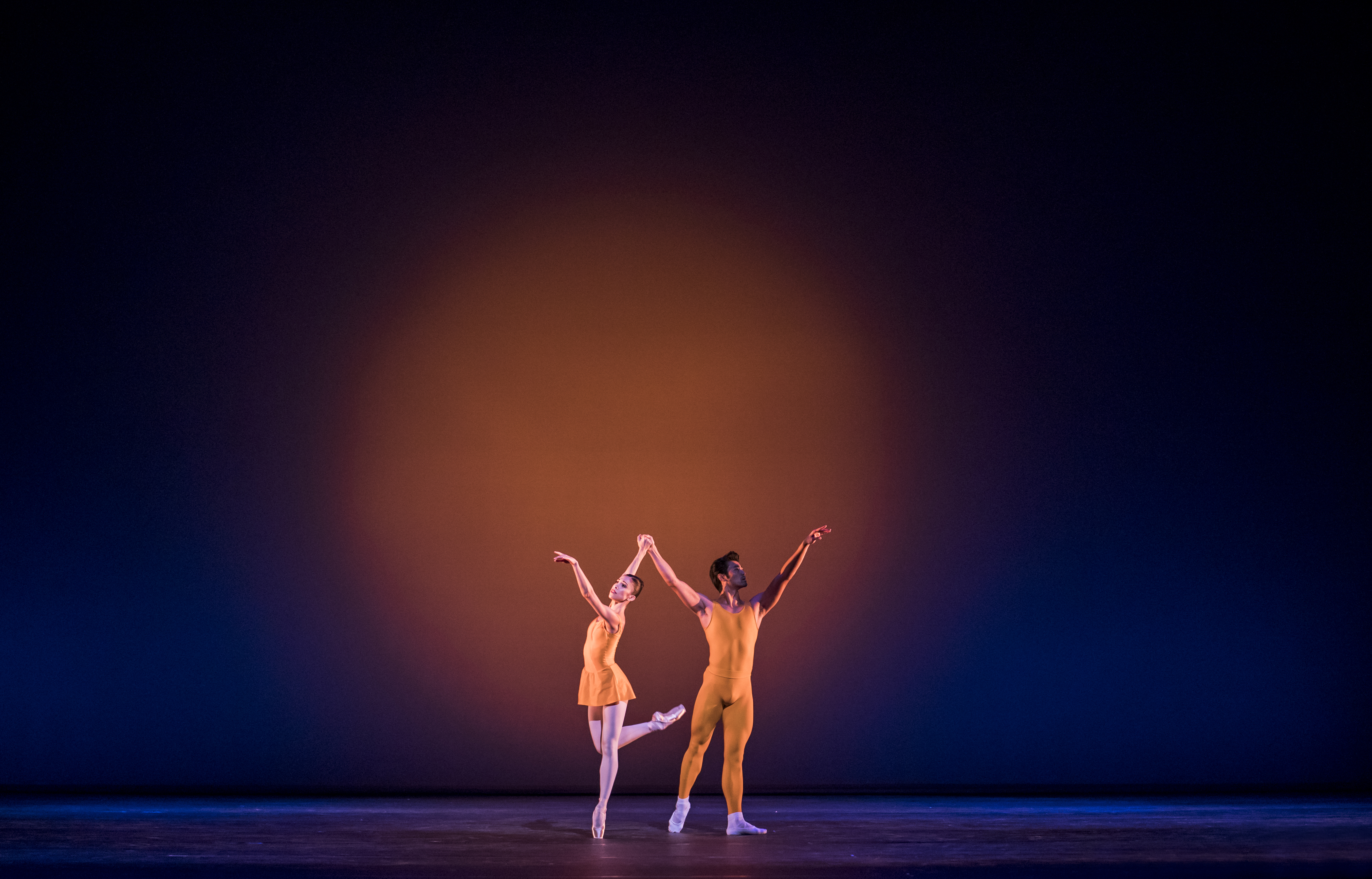 Two dancers wearing yellow leotards appear on stage. The backdrop is lit with a blue light, with a yellow spotlight in the centre. The dancers stand arms raised with their hands joined. The dancer on the left has her leg raised.