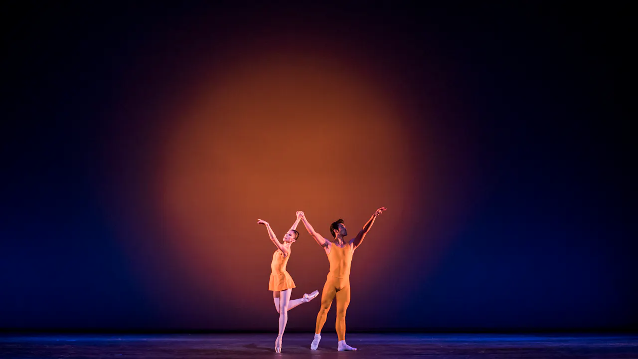 Two dancers wearing yellow leotards appear on stage. The backdrop is lit with a blue light, with a yellow spotlight in the centre. The dancers stand arms raised with their hands joined. The dancer on the left has her leg raised.