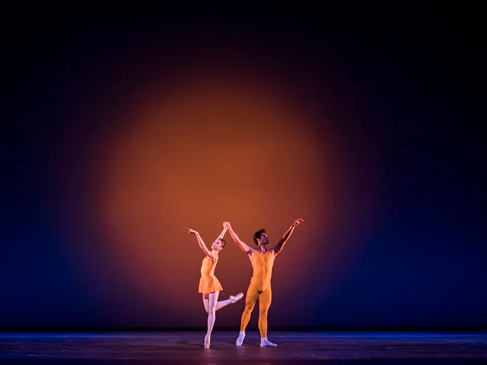 Two dancers wearing yellow leotards appear on stage. The backdrop is lit with a blue light, with a yellow spotlight in the centre. The dancers stand arms raised with their hands joined. The dancer on the left has her leg raised.