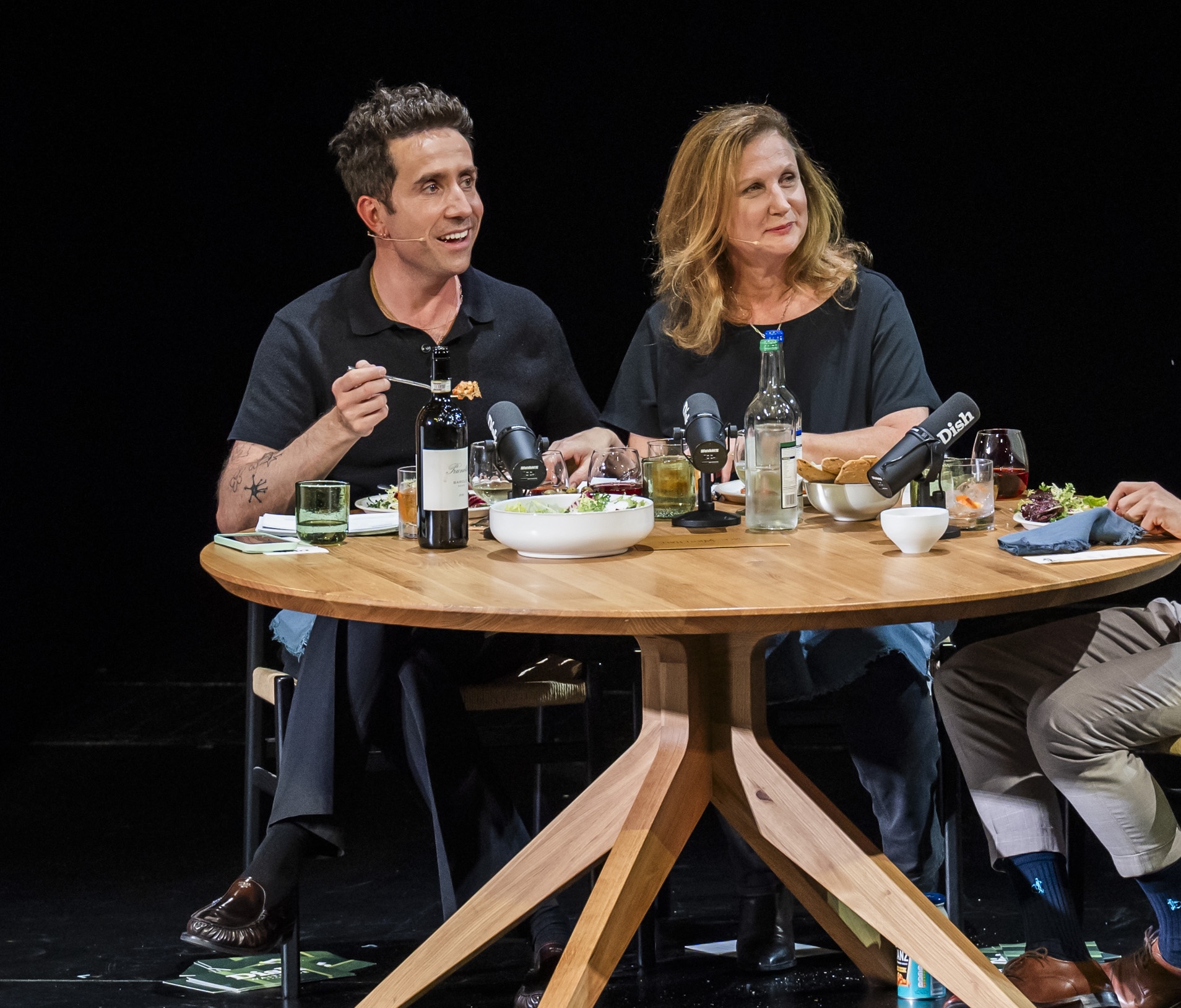 Angela Harnett and Nick Grimshaw sit at a wooden table set with various plates of food and bottles of wine. 