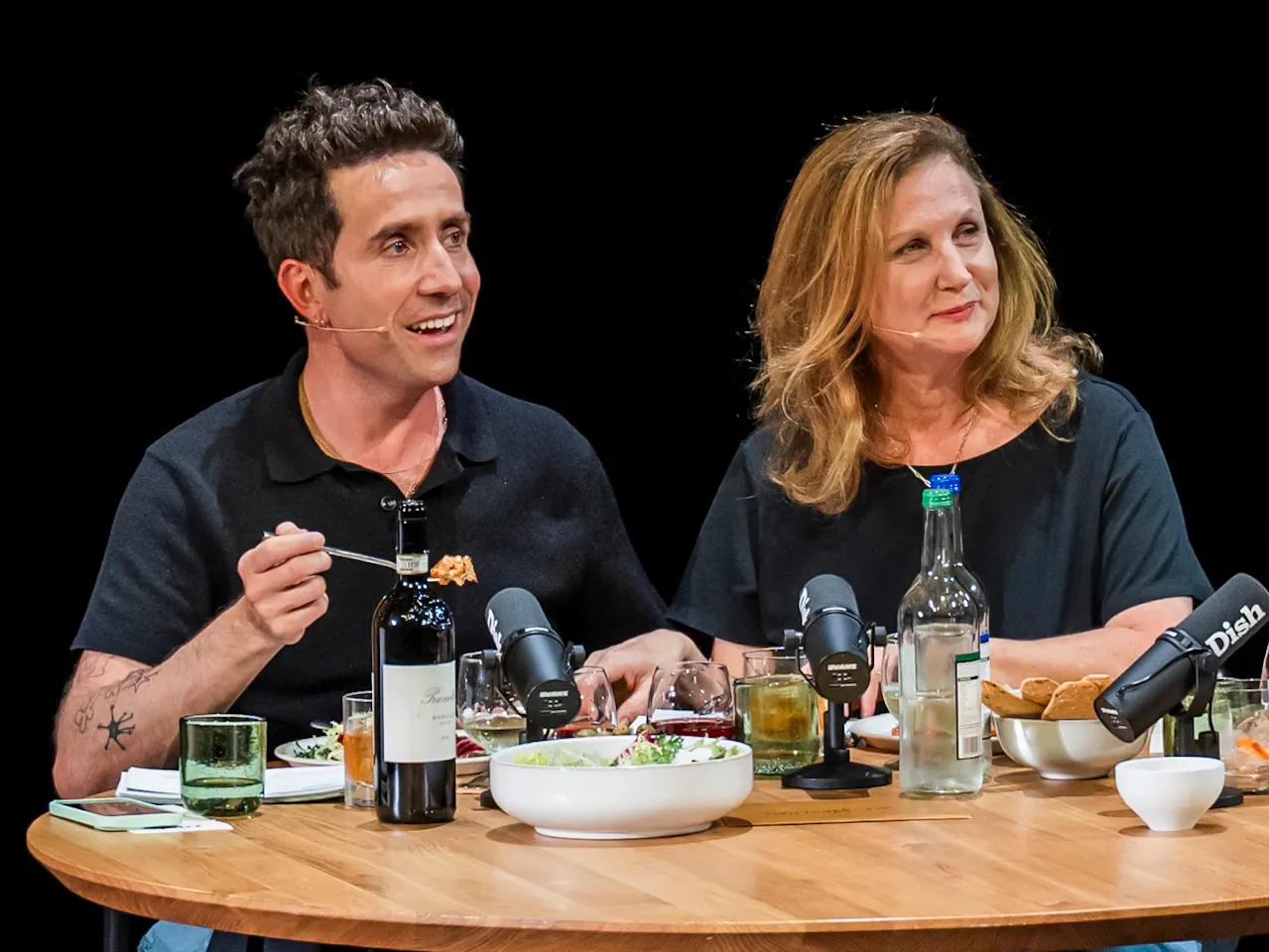 Angela Harnett and Nick Grimshaw sit at a wooden table set with various plates of food and bottles of wine.