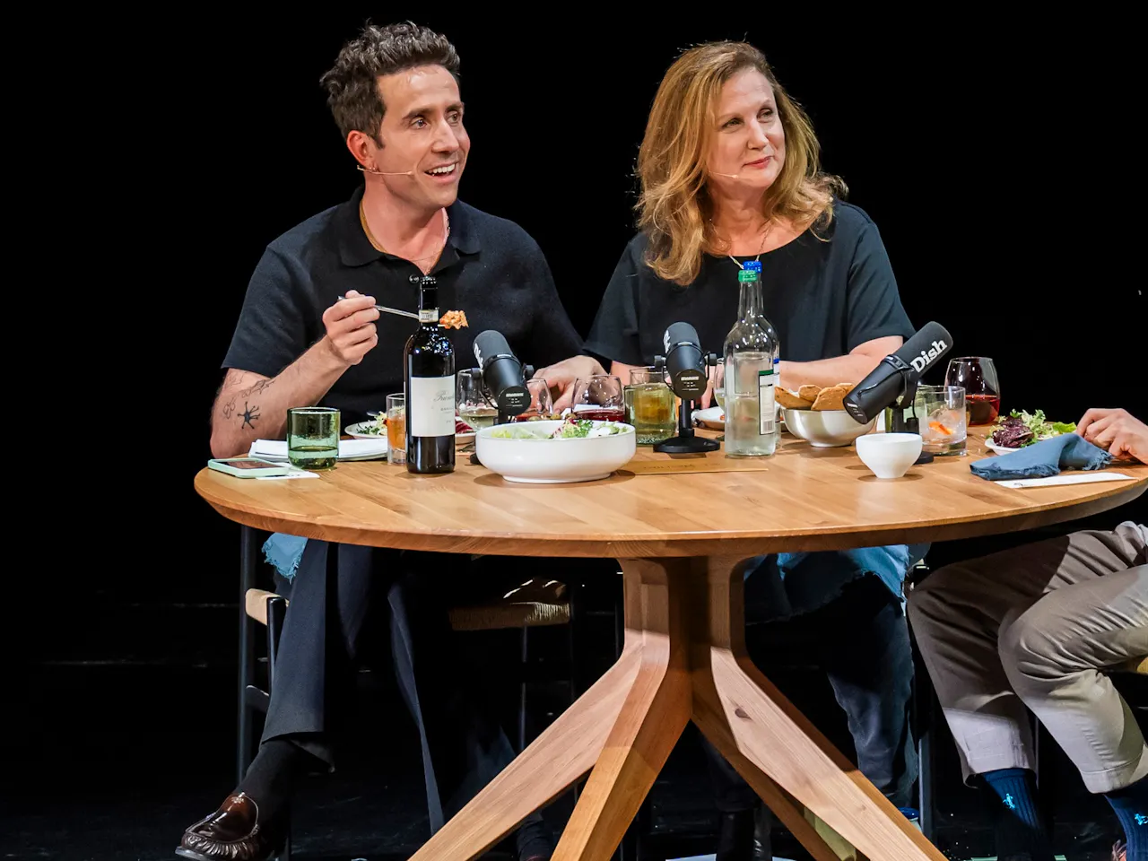 Angela Harnett and Nick Grimshaw sit at a wooden table set with various plates of food and bottles of wine.