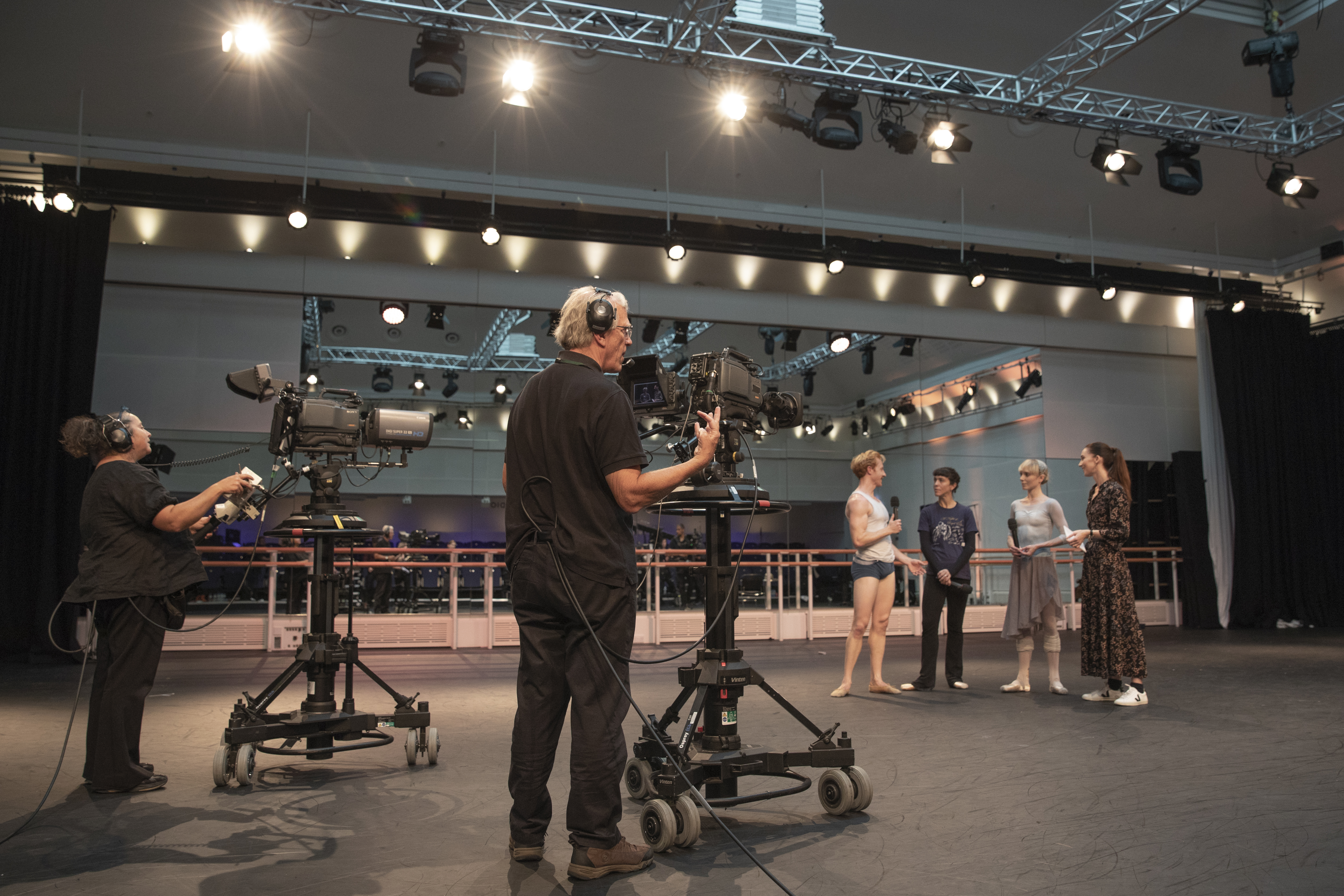 A group of people stand in conversation in the middle of a ballet studio, two cameras are pointed at them. (3 of 8)