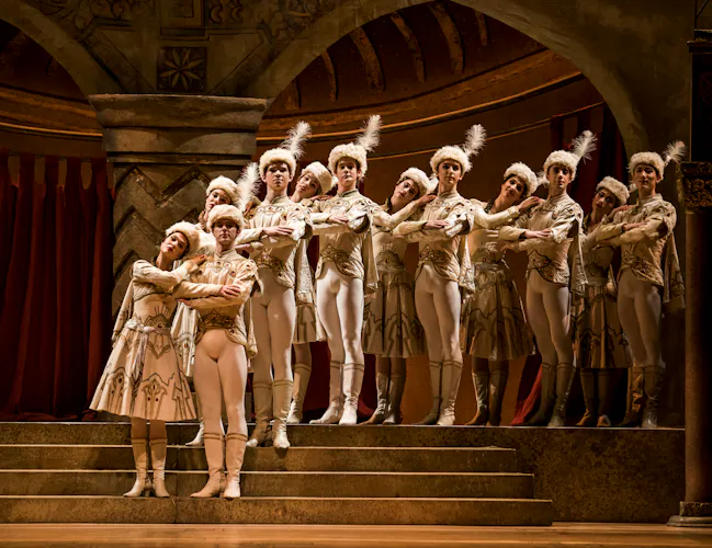 A group of dancers stand on a set of steps on stage, behind them are ornate archways. They wear decorative formal white outfits and have fur hats with white feathers. (2 of 3)