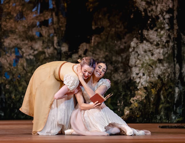 Yasmine Naghdi and Anna Rose O'Sullivan performing as Tatiana and Olga. Tatiana holds Olga's cheek to her face in a moment of sisterly affection, a book in her other hand.