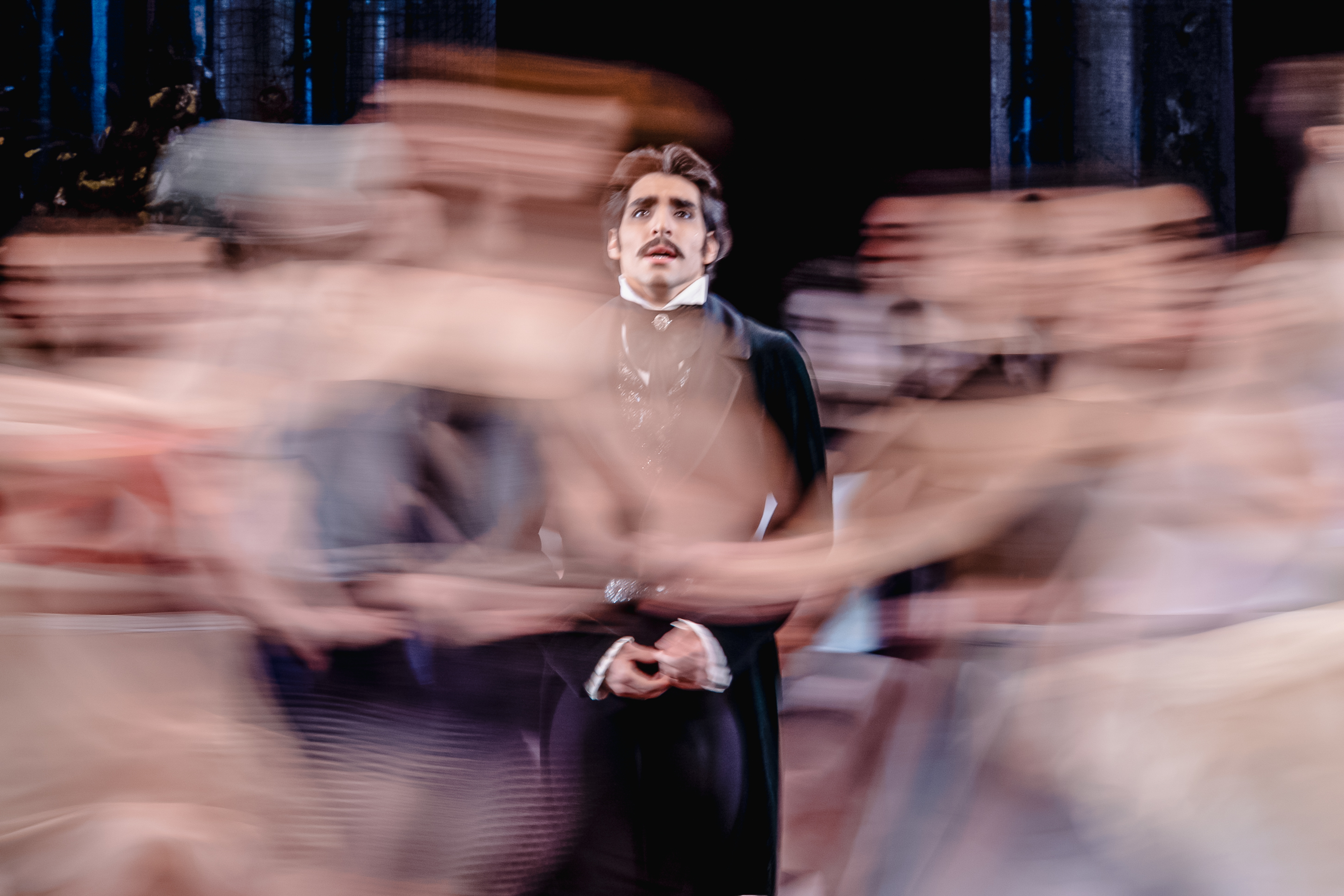 Cesar Corrales performing as Eugene Onegin. Dressed in a black suit, he is staring upwards as a swirl of commotion surrounds him. 