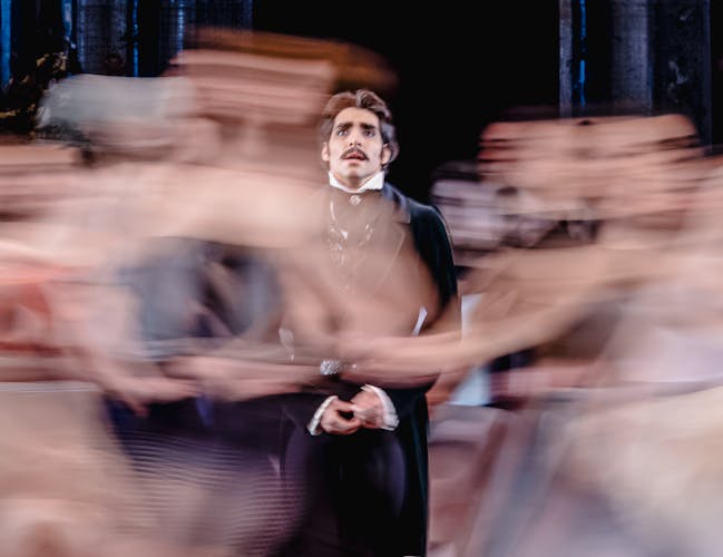 Cesar Corrales performing as Eugene Onegin. Dressed in a black suit, he is staring upwards as a swirl of commotion surrounds him.