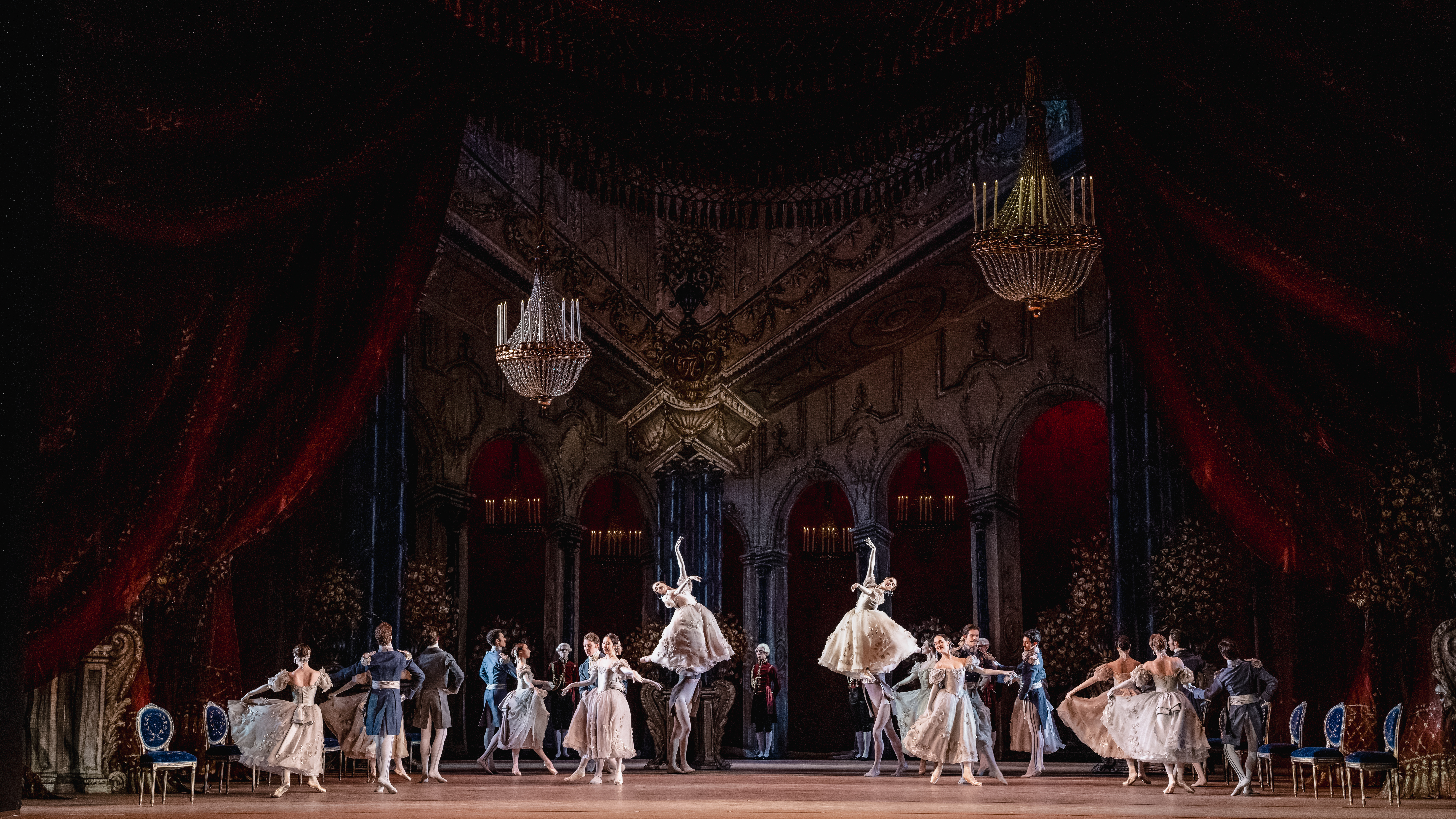 Dancers waltz together in pairs throughout the ballroom, some women are lifted high in the air by their partners. 