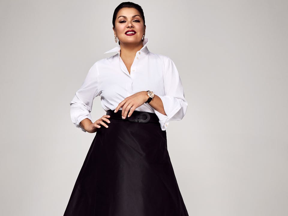 The opera singer Anna Netrebko stands against a white wall in a photography studio wearing a white shirt, long black skirt and black high heels. She smiles towards the camera.