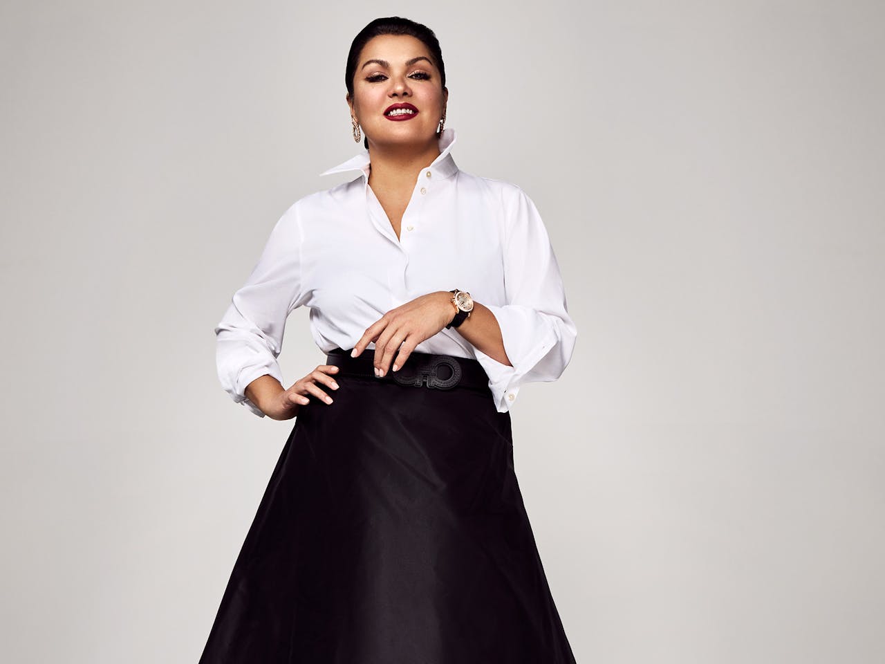 The opera singer Anna Netrebko stands against a white wall in a photography studio wearing a white shirt, long black skirt and black high heels. She smiles towards the camera.