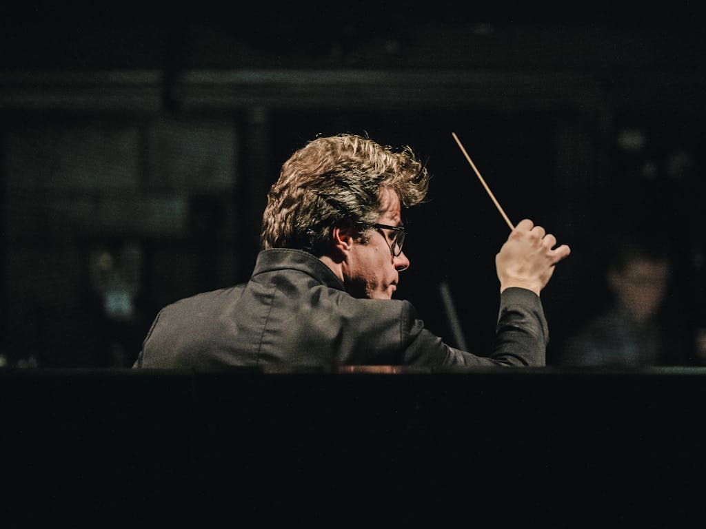 The music Director of The Royal Opera, Jakub Hrůša wears a black jacket and black rimmed glasses as he stands in an orchestra pit of The Royal Opera House with his back against the audience. Members of the orchestra are slightly blurred but visible in front of him. He holds a baton up in his right hand.