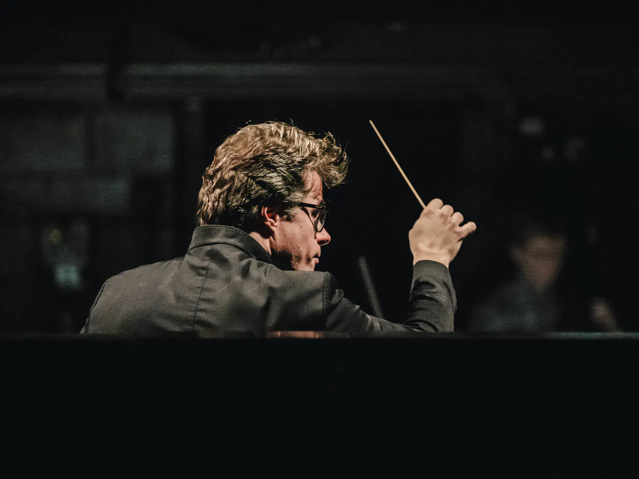 The music Director of The Royal Opera, Jakub Hrůša wears a black jacket and black rimmed glasses as he stands in an orchestra pit of The Royal Opera House with his back against the audience. Members of the orchestra are slightly blurred but visible in front of him. He holds a baton up in his right hand.
