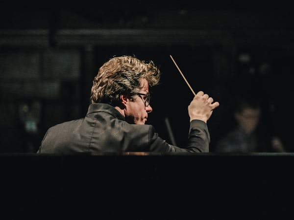 The music Director of The Royal Opera, Jakub Hrůša wears a black jacket and black rimmed glasses as he stands in an orchestra pit of The Royal Opera House with his back against the audience. Members of the orchestra are slightly blurred but visible in front of him. He holds a baton up in his right hand.