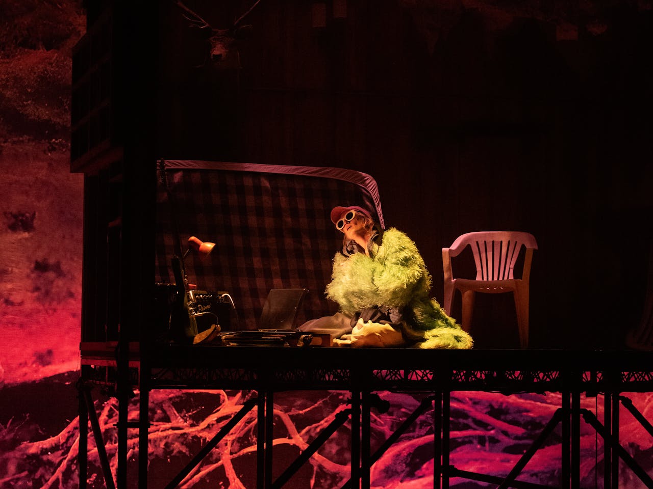 A performer sits on a bent mattress on the floor wearing a fake fur coat, baseball cap and white rimmed cat-eye style sunglasses. Next to them is a white plastic garden chair and above them the a small square of ceiling appears to be made from corrugated plastic. The stage is lit with pink lighting but golden lighting shines up from the performer making them appear to be in bright yellow light.