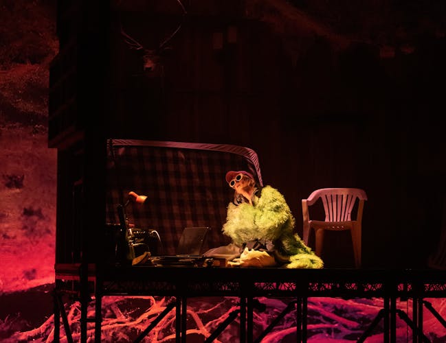 A performer sits on a bent mattress on the floor wearing a fake fur coat, baseball cap and white rimmed cat-eye style sunglasses. Next to them is a white plastic garden chair and above them the a small square of ceiling appears to be made from corrugated plastic. The stage is lit with pink lighting but golden lighting shines up from the performer making them appear to be in bright yellow light.