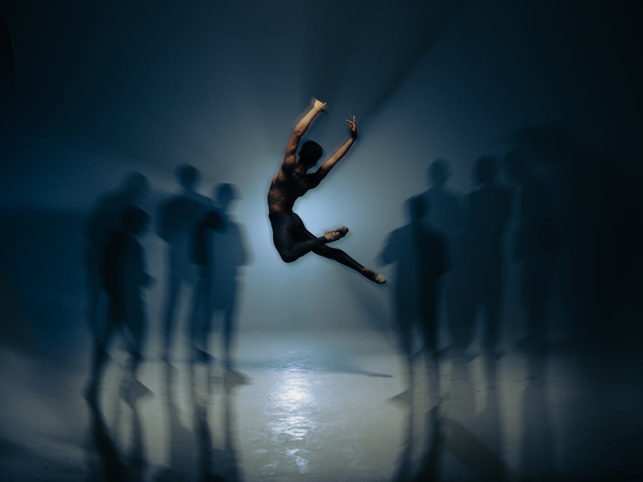 A ballet dancer with short black hair wearing leggings and ballet slippers launches themselves into the air - they are only visible from behind as those arms are raised above their shoulders, their left leg bent and their right leg stretched outwards making a shape similar to the letter C. In front of them are the blurry shadows of around 8 or 9 dancers but as the stage is set dark with only light on the dancer in the centre, they are impossible to decipher making them almost translucent.