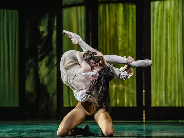 The ballet dancer Matthew Ball is performing as Crown Prince Rudolf in Mayerling. He kneels on the floor of the stage with green fabric lined folding doors behind him. In his arms he holds up the ballet dancer Melissa Hamilton, who is performing as Mary Vetsera. He kisses her lips, as her legs wrap around his back and neck. Their bodies are contorted to create a T-shape.