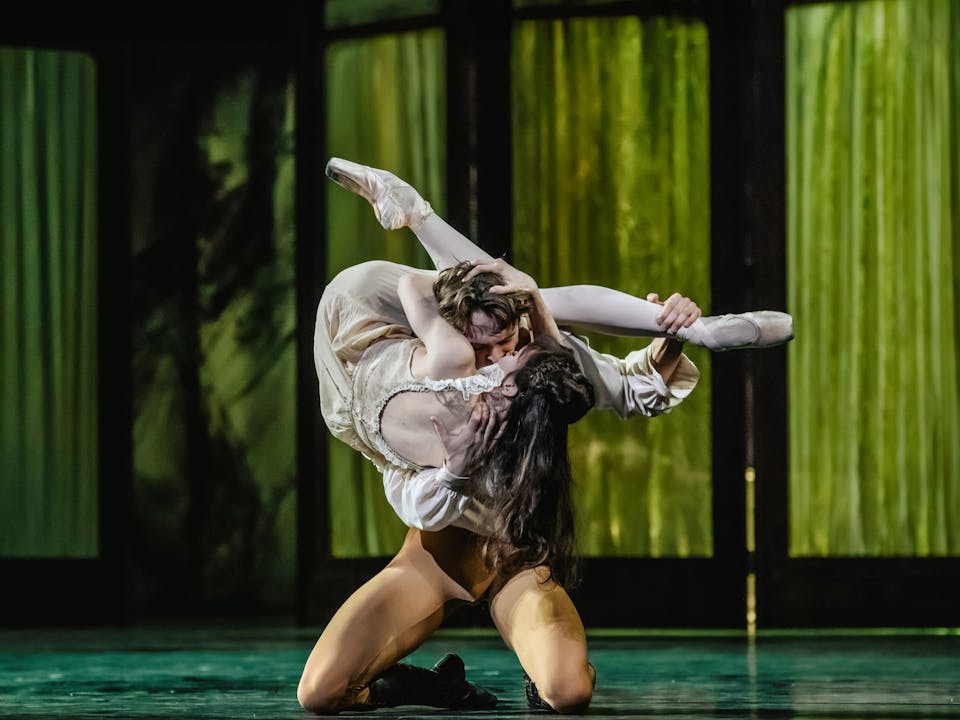 The ballet dancer Matthew Ball is performing as Crown Prince Rudolf in Mayerling. He kneels on the floor of the stage with green fabric lined folding doors behind him. In his arms he holds up the ballet dancer Melissa Hamilton, who is performing as Mary Vetsera. He kisses her lips, as her legs wrap around his back and neck. Their bodies are contorted to create a T-shape.