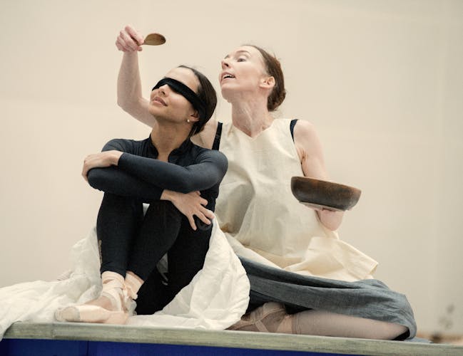 The Royal Ballet dancers Francesca Hayward and Kristen McNally are together in a rehearsal studio rehearsing Like Water for Chocolate. Francesca Hayward wears all black, pink ballet shoes. a white skirt over the black leggings and a black mask over her eyes. She sits on her skirt with her arms crossed over her chest and looks upwards. Behind her, Kisten McNally sits holding up a wooden bowl in her left hand and a wooden spoon in her right hand above Haywards head